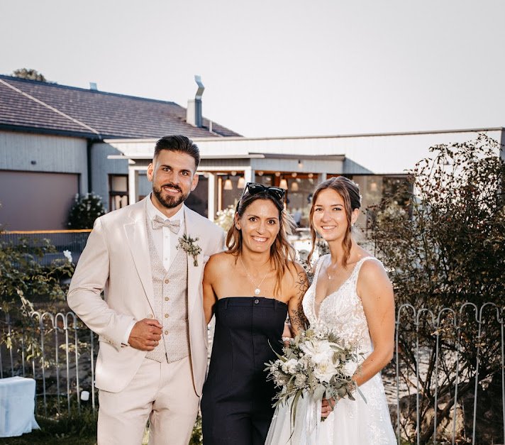Drei Menschen bei einer Hochzeit, zwei Frauen und ein Mann, im Freien, im Hintergrund ein modernes Haus, die Frau in einem weißen Brautkleid hält einen Blumenstrauß.