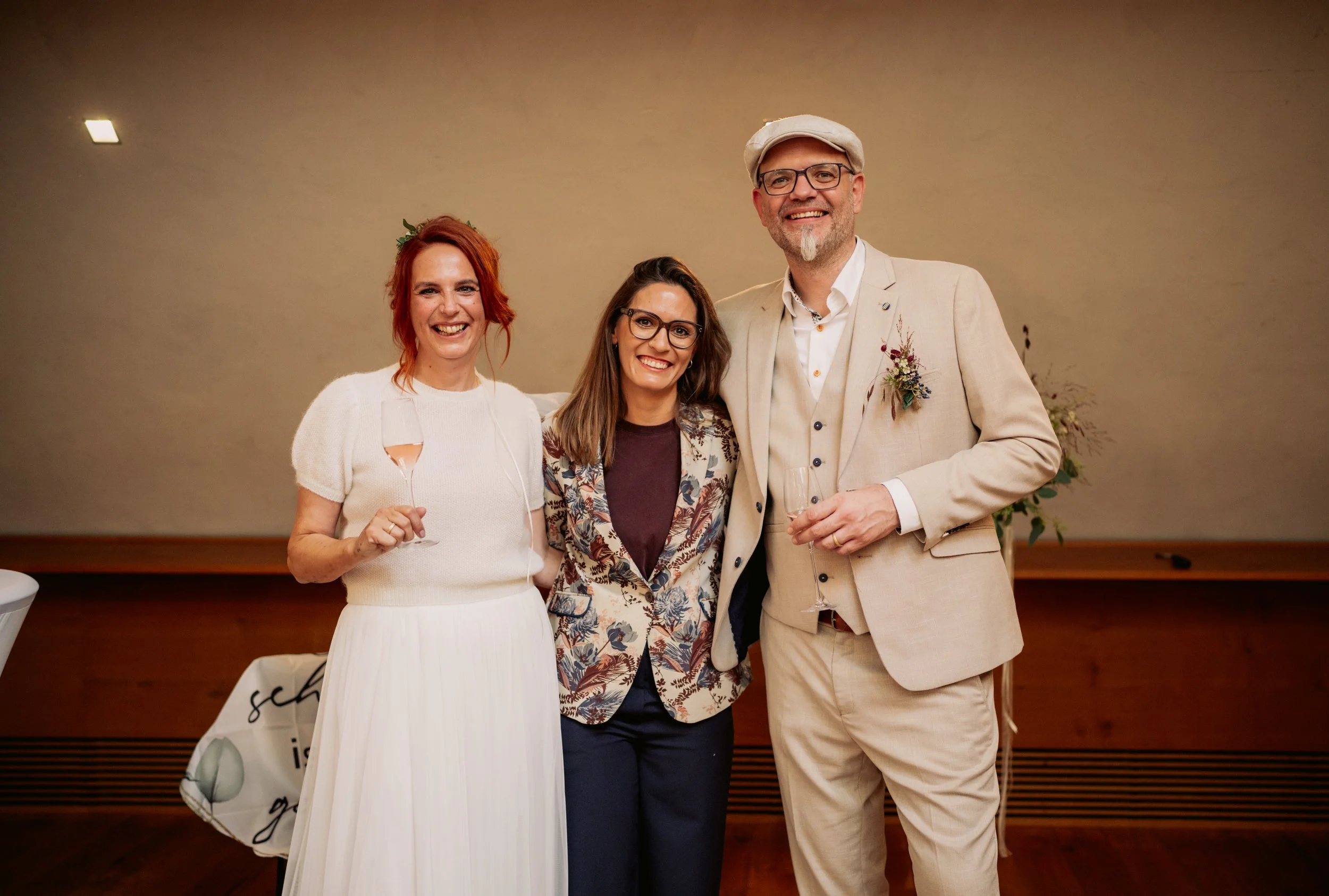 Drei Menschen bei einer Feier, alle lächelnd, mit Getränken in der Hand, im Hintergrund eine einfache Wand und eine größere Pflanze.