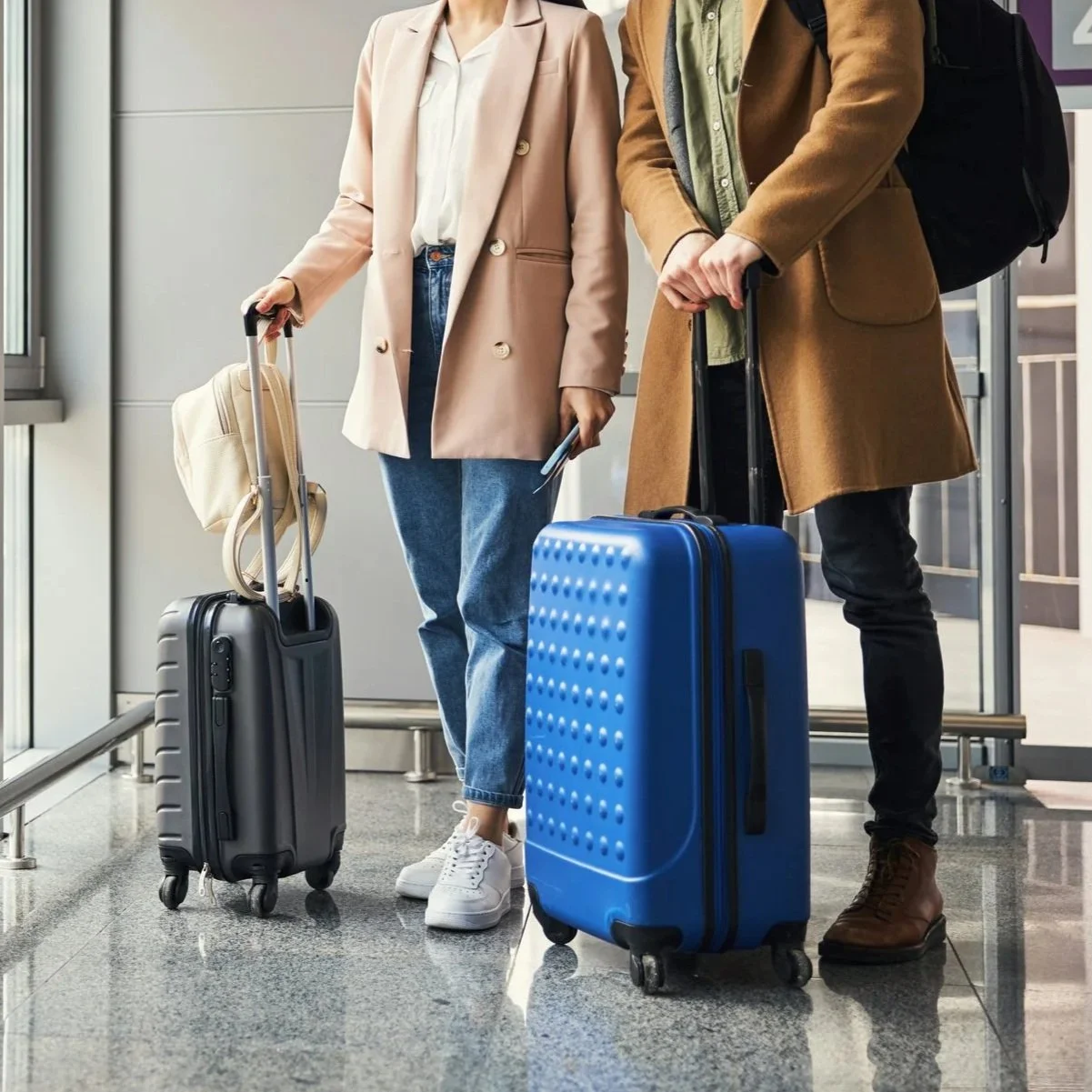 Two people standing with luggage at an airport, one with a black suitcase and another with a blue suitcase.