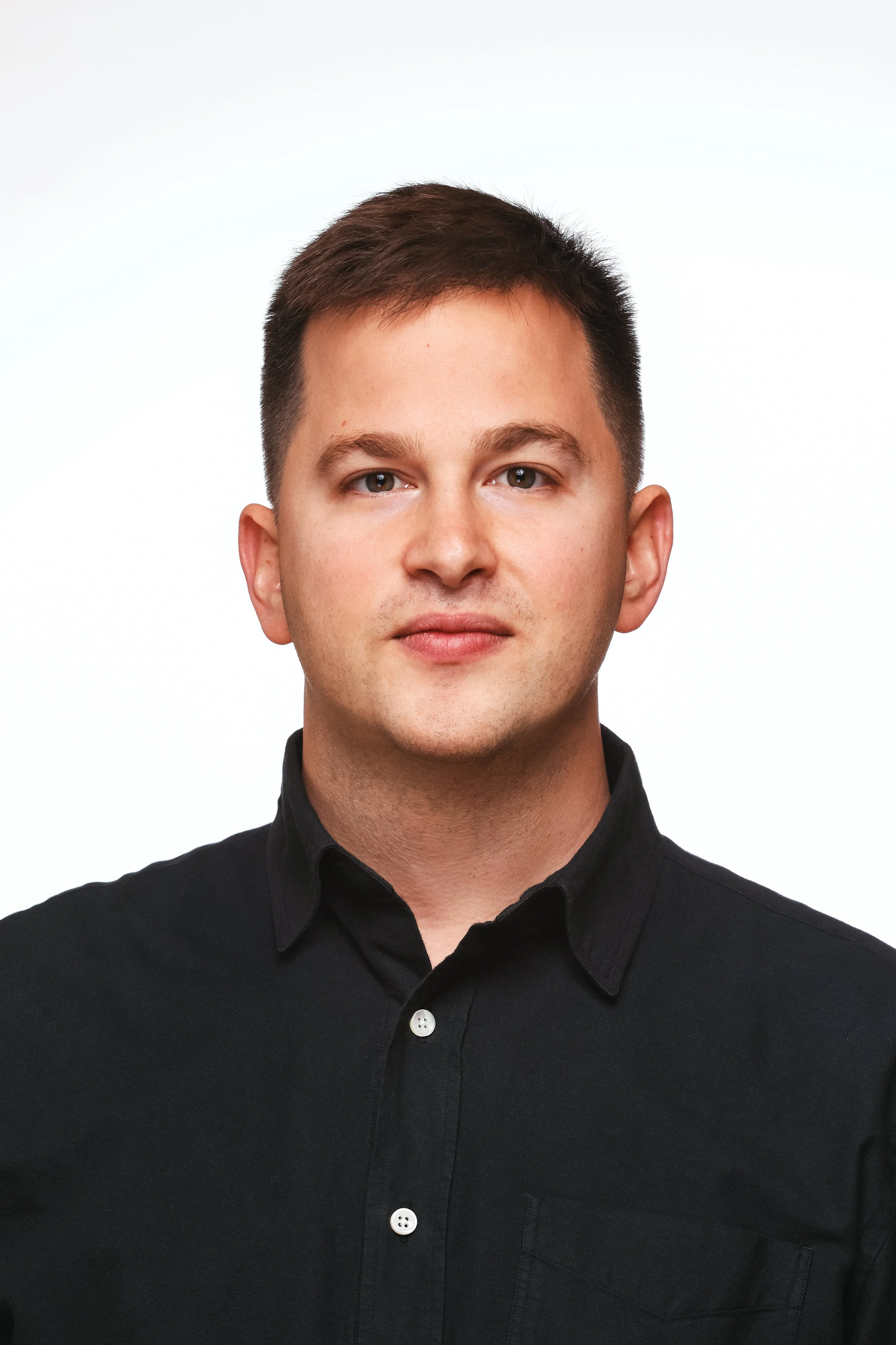 A young man with short brown hair, wearing a black button-up shirt, facing the camera against a plain white background.
