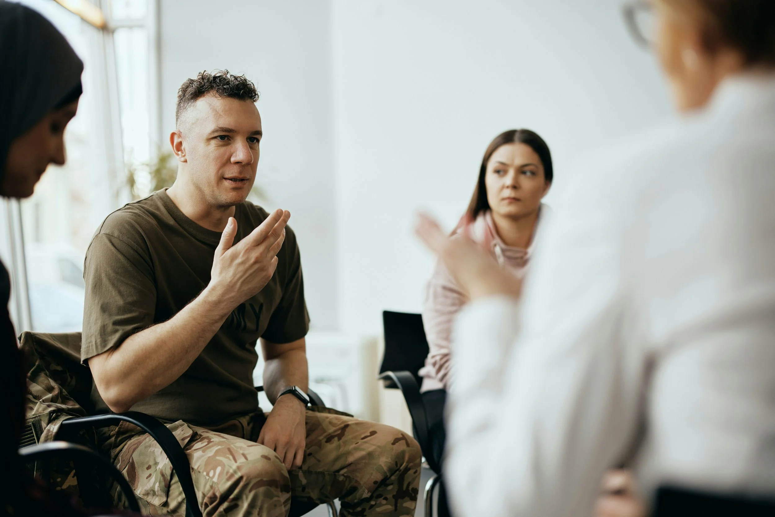 A man in military camouflage pants and a brown t-shirt gestures while talking to a group of people in a discussion room.