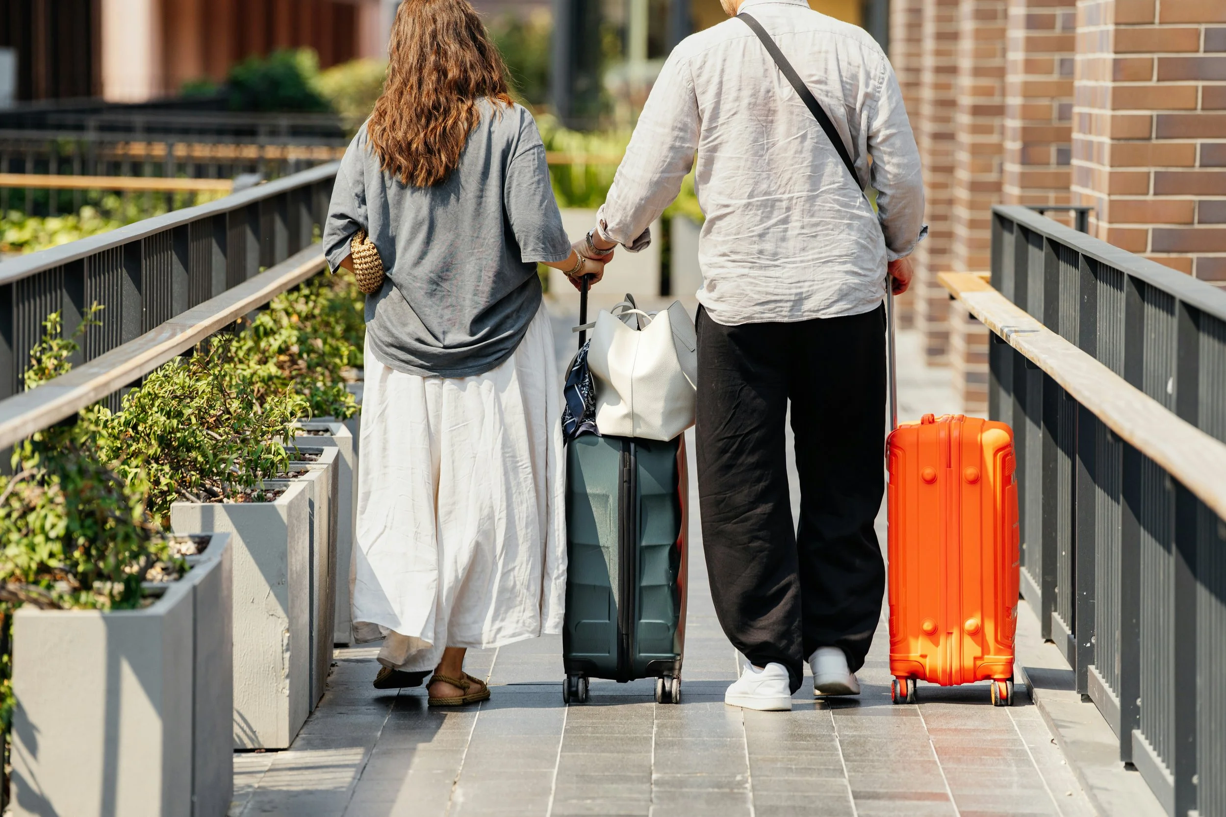 A couple walking on a pedestrian bridge with luggage, holding hands, with planters and a brick building in the background.