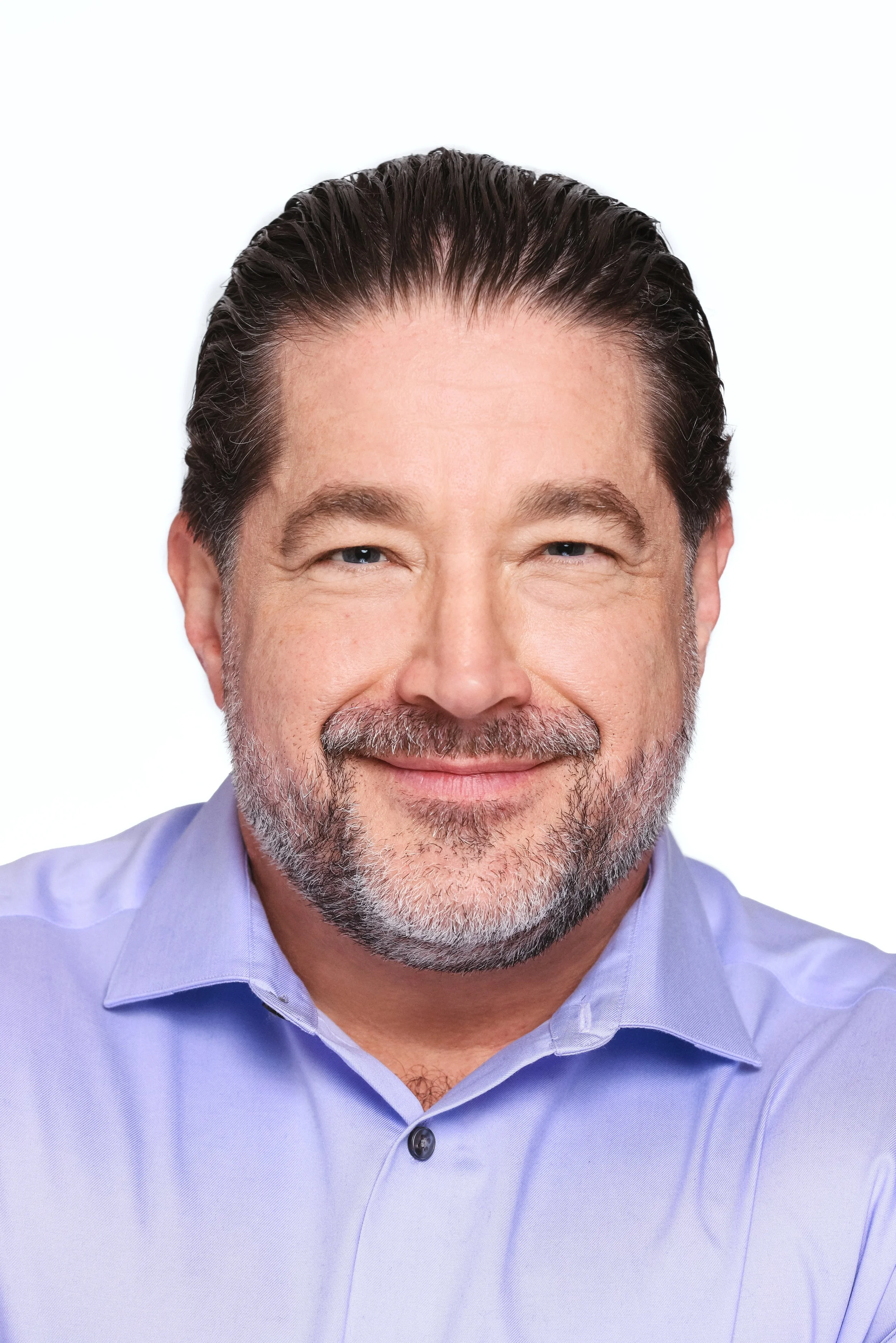 Close-up image of a smiling middle-aged man with dark hair, a beard with gray, wearing a light blue dress shirt against a plain white background.