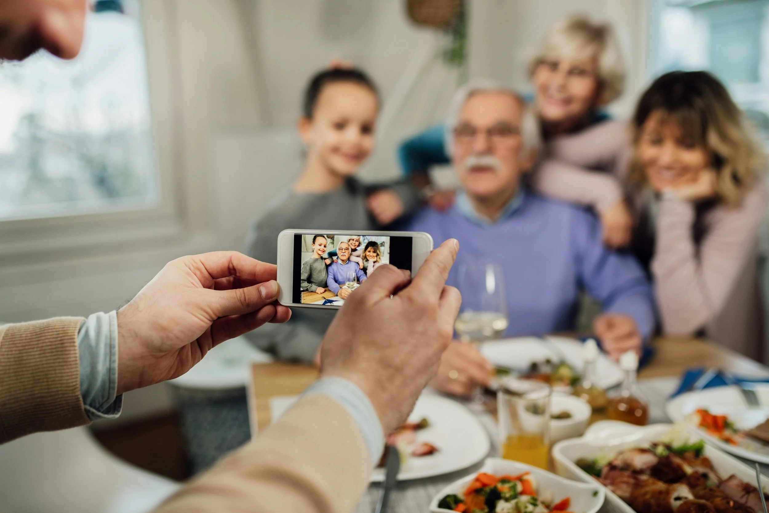Person taking a photo of a family gathered around a table with food, with the family seen on the phone screen.