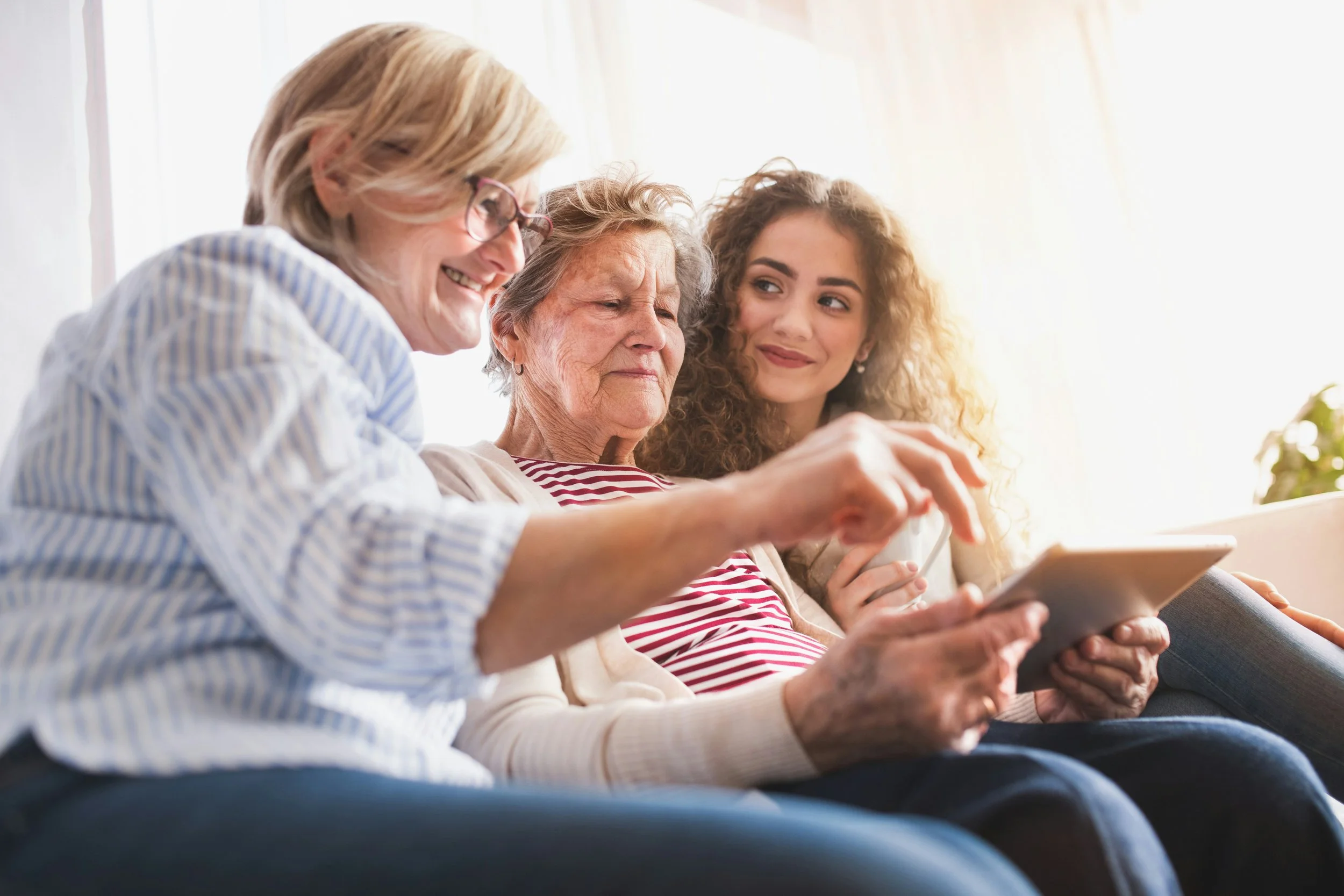 Three women sitting on a sofa, looking at a tablet and smiling.