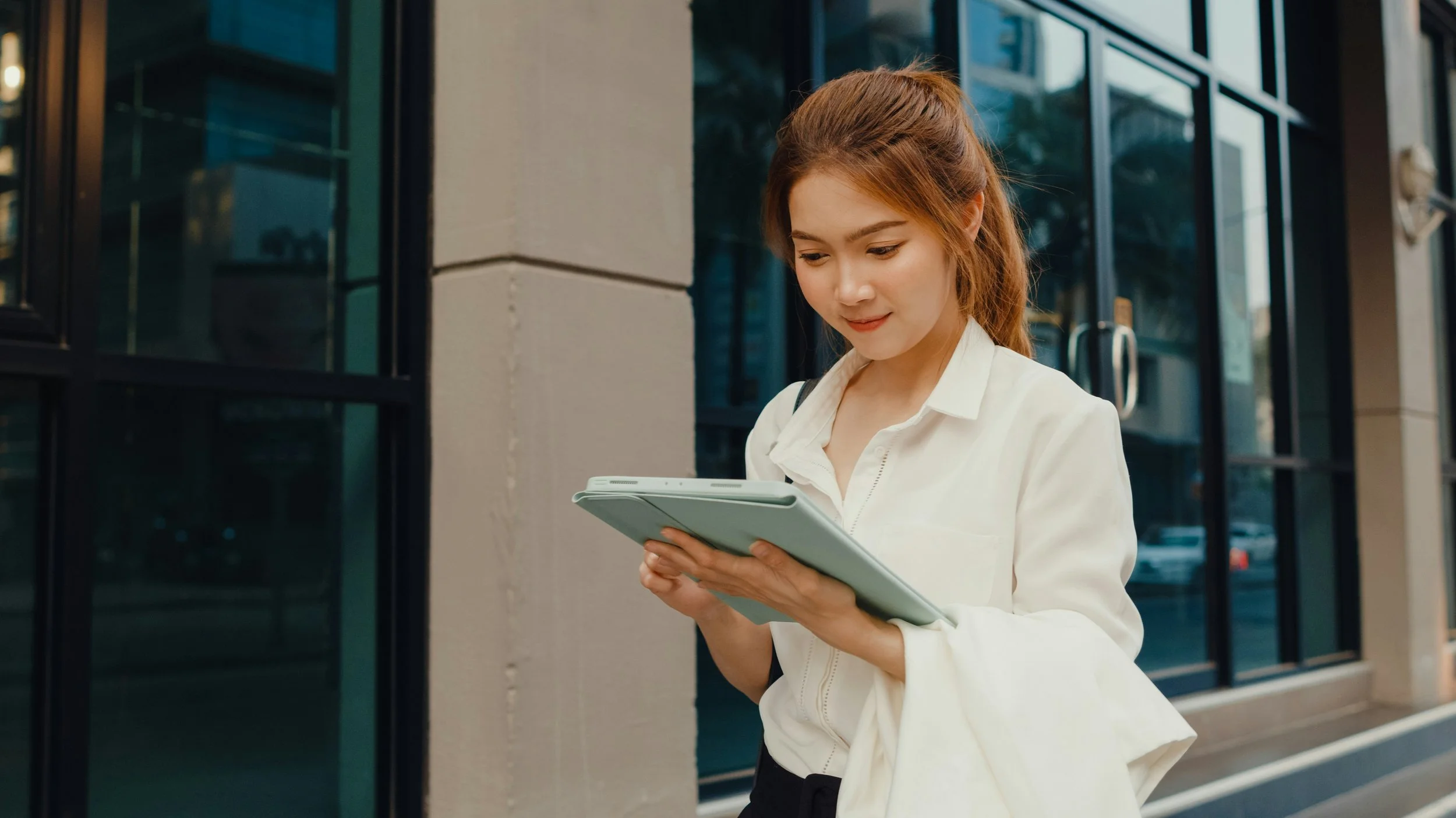 Young woman with red hair reading a tablet outside a building with large glass windows.