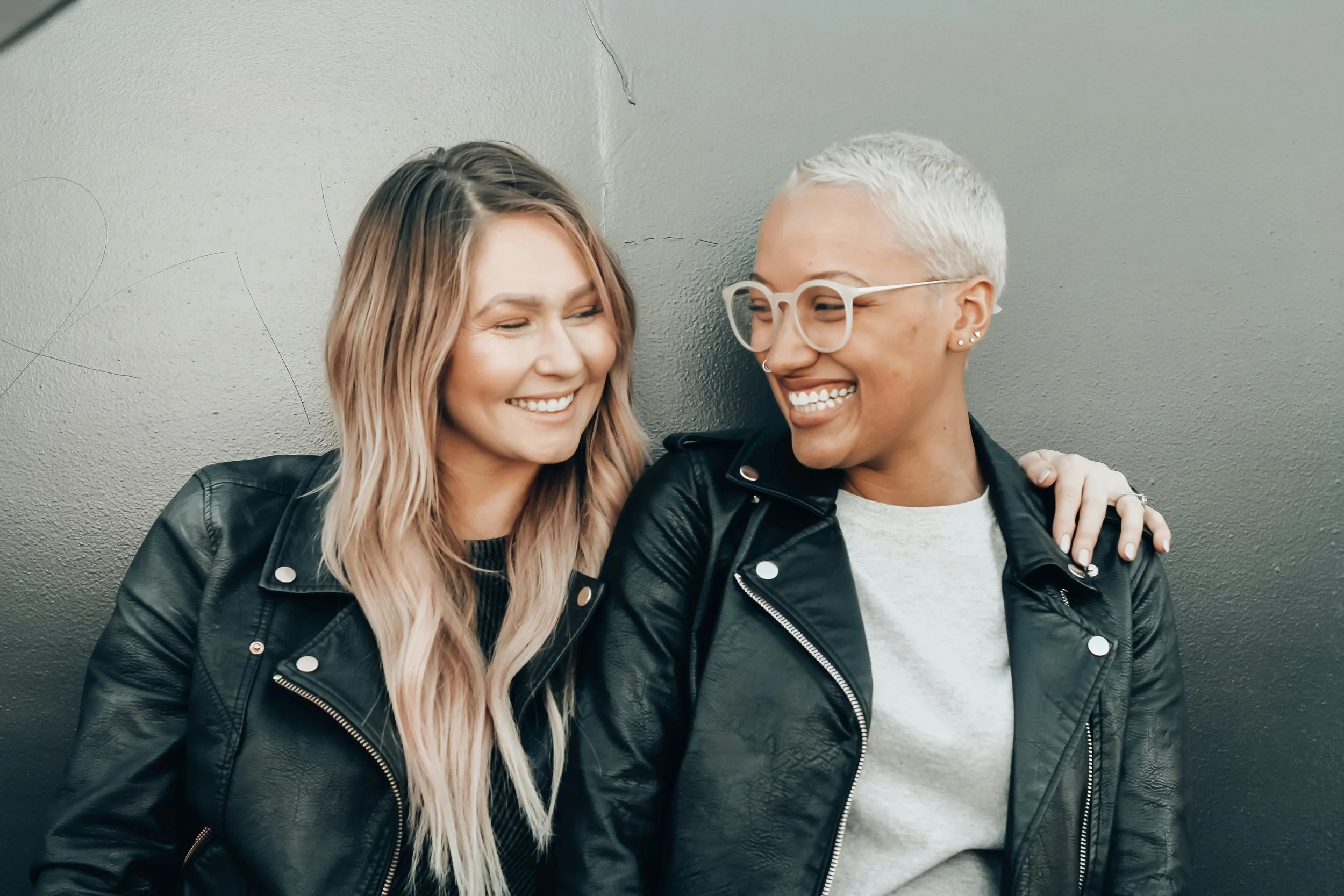 Two women smiling and sharing an affectionate moment, leaning against a gray wall, both wearing black leather jackets.