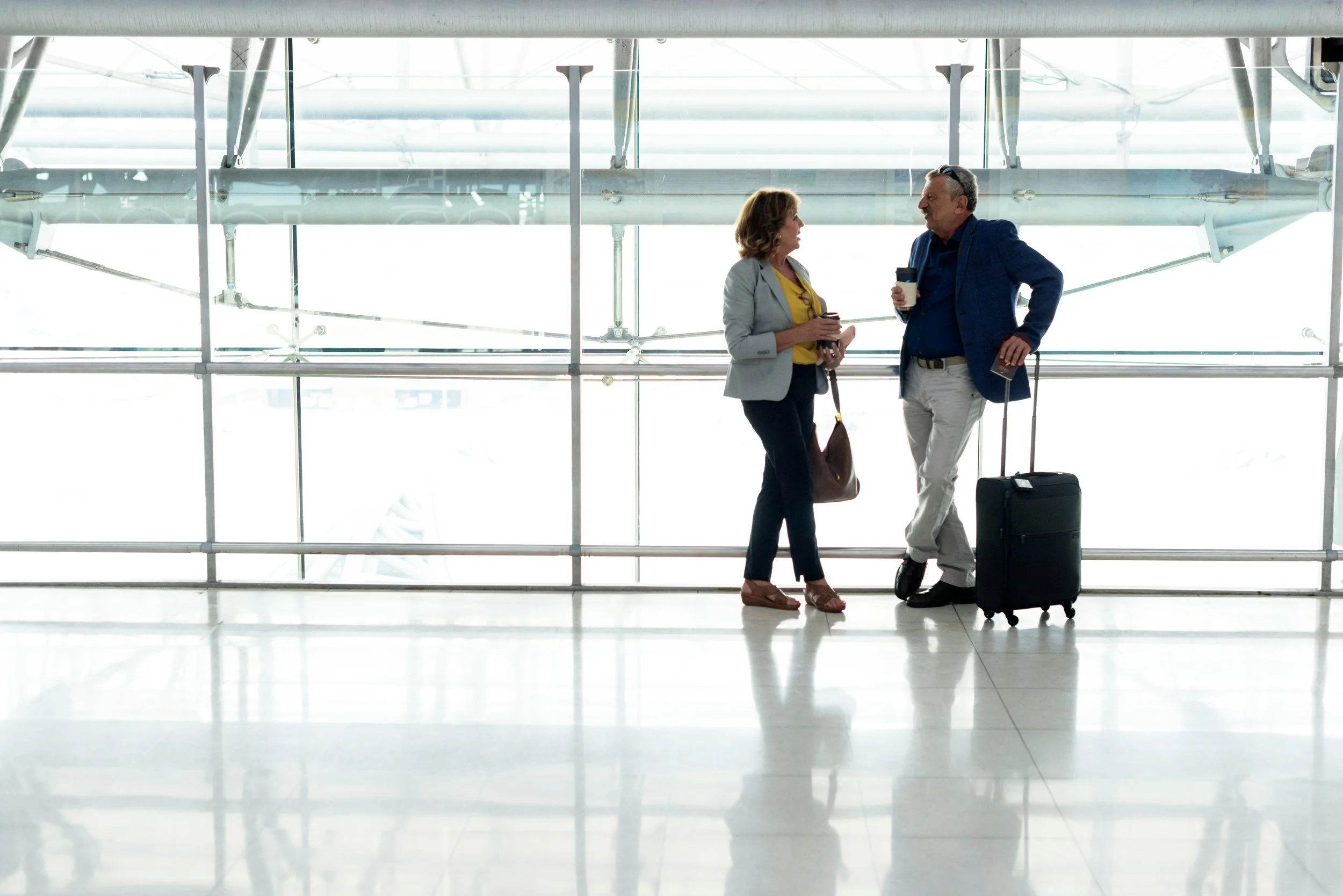 Two adults, a man and a woman, standing in an airport terminal near large windows, having a conversation. The woman holds a smartphone and a coffee cup, and carries a handbag. The man holds a coffee cup and has a rolling suitcase.