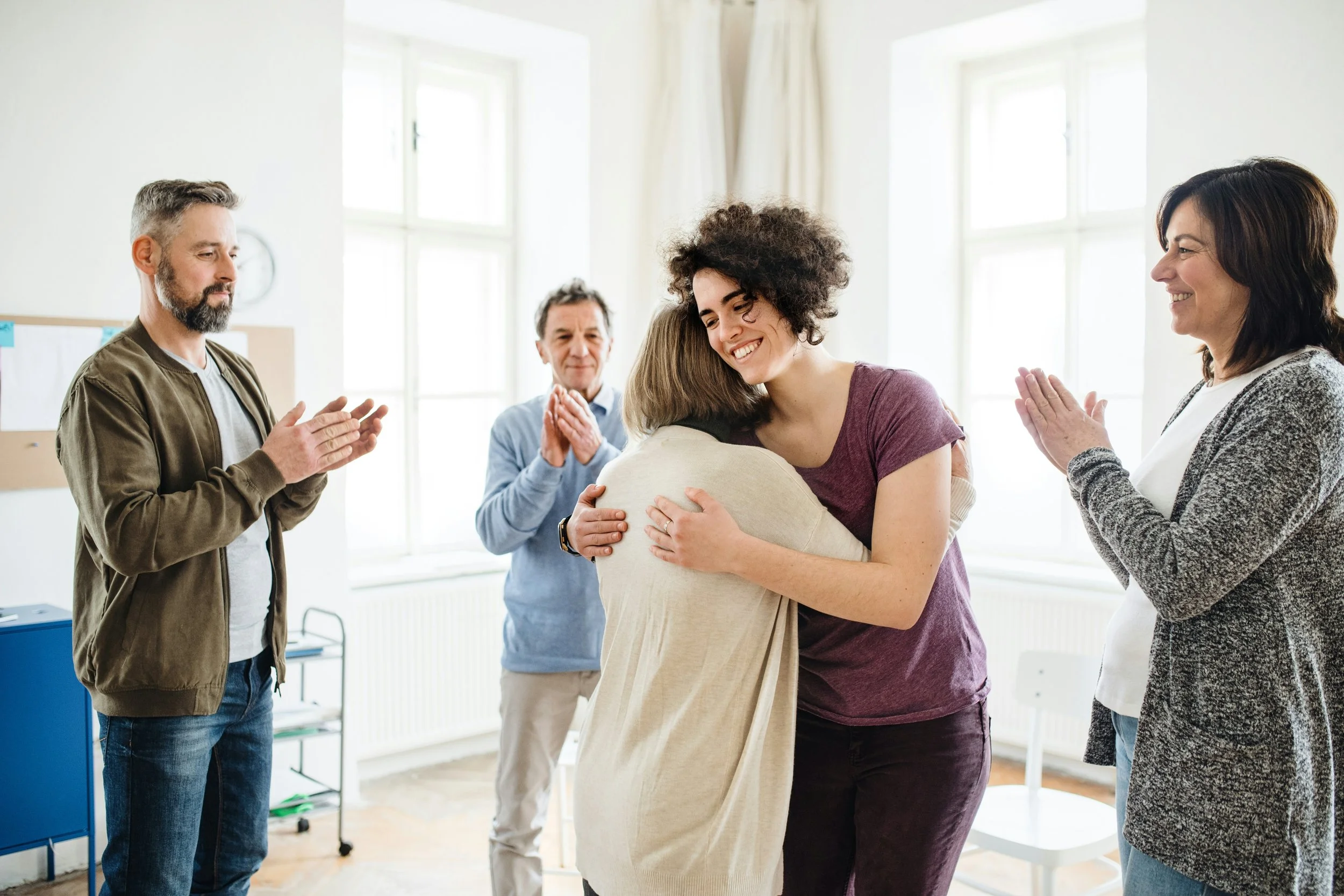 A diverse group of five people in a bright room with large windows, hugging and clapping, celebrating a positive moment.