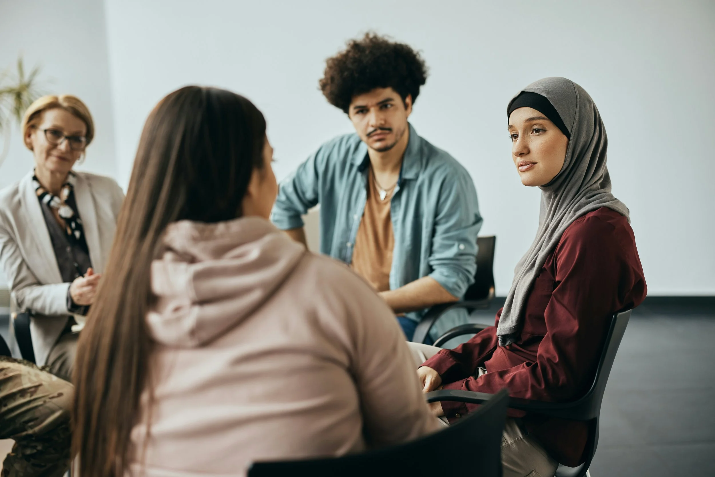 A diverse group of women and a man having a discussion in a meeting room, with one woman wearing a hijab and a woman in camouflage pants.