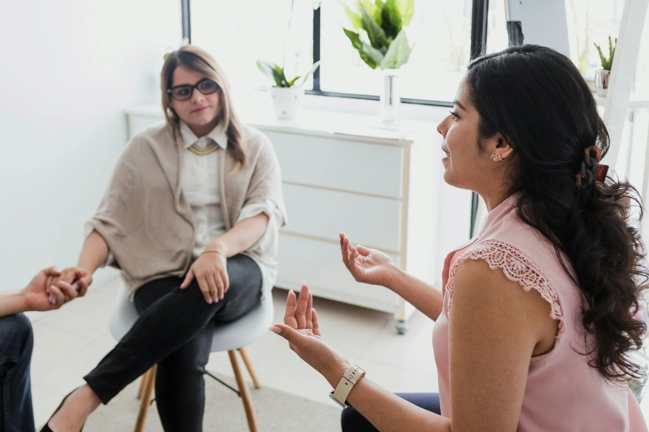 Two women sitting in a bright room, engaged in a conversation. One woman, with dark curly hair, wearing a pink sleeveless top, is gesturing with her hands. The other woman, with straight hair and glasses, dressed in a beige cardigan and white shirt, is sitting on a chair.