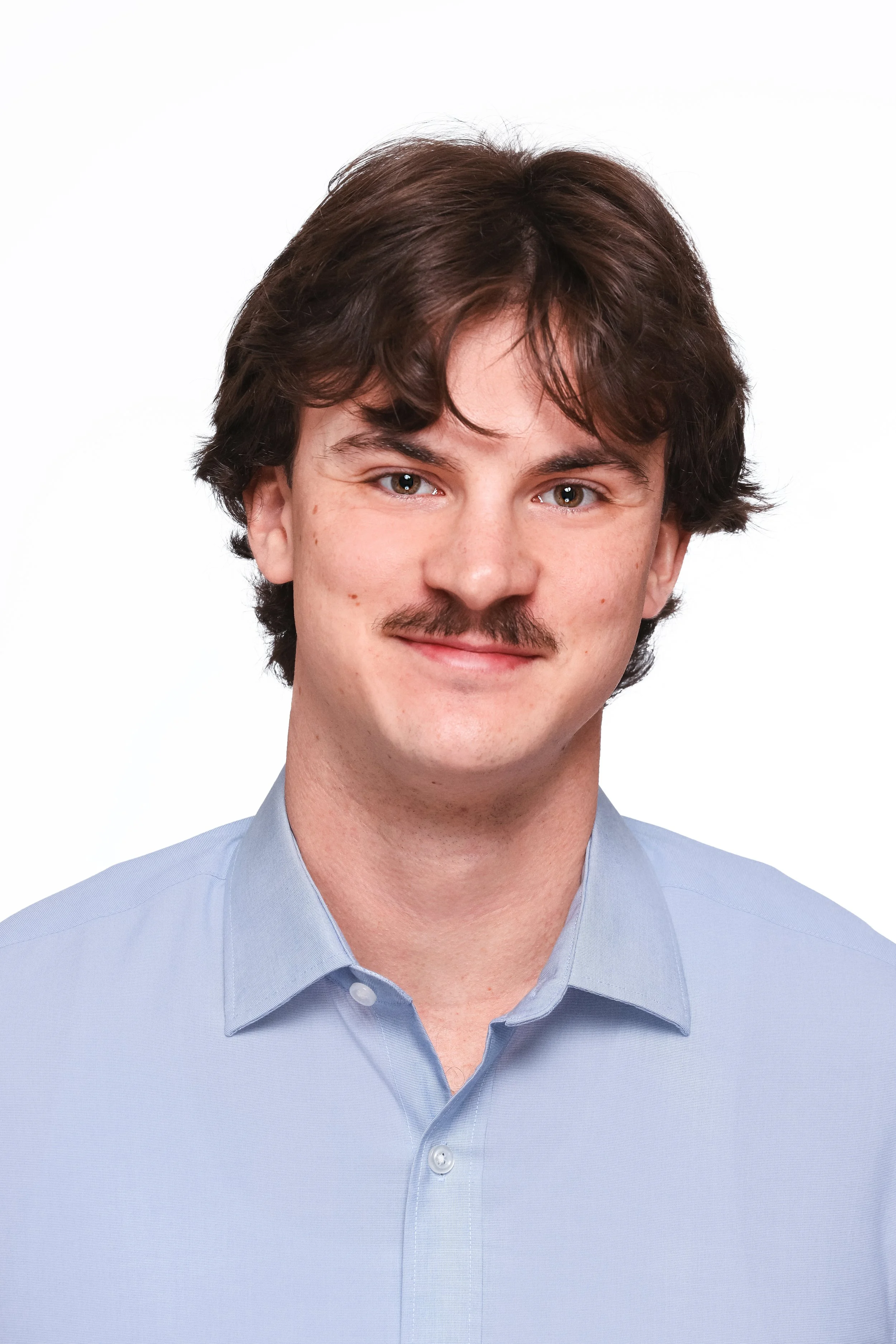 Headshot of a young man with brown hair, mustache, blue eyes, and light skin, wearing a light blue collared shirt against a white background.
