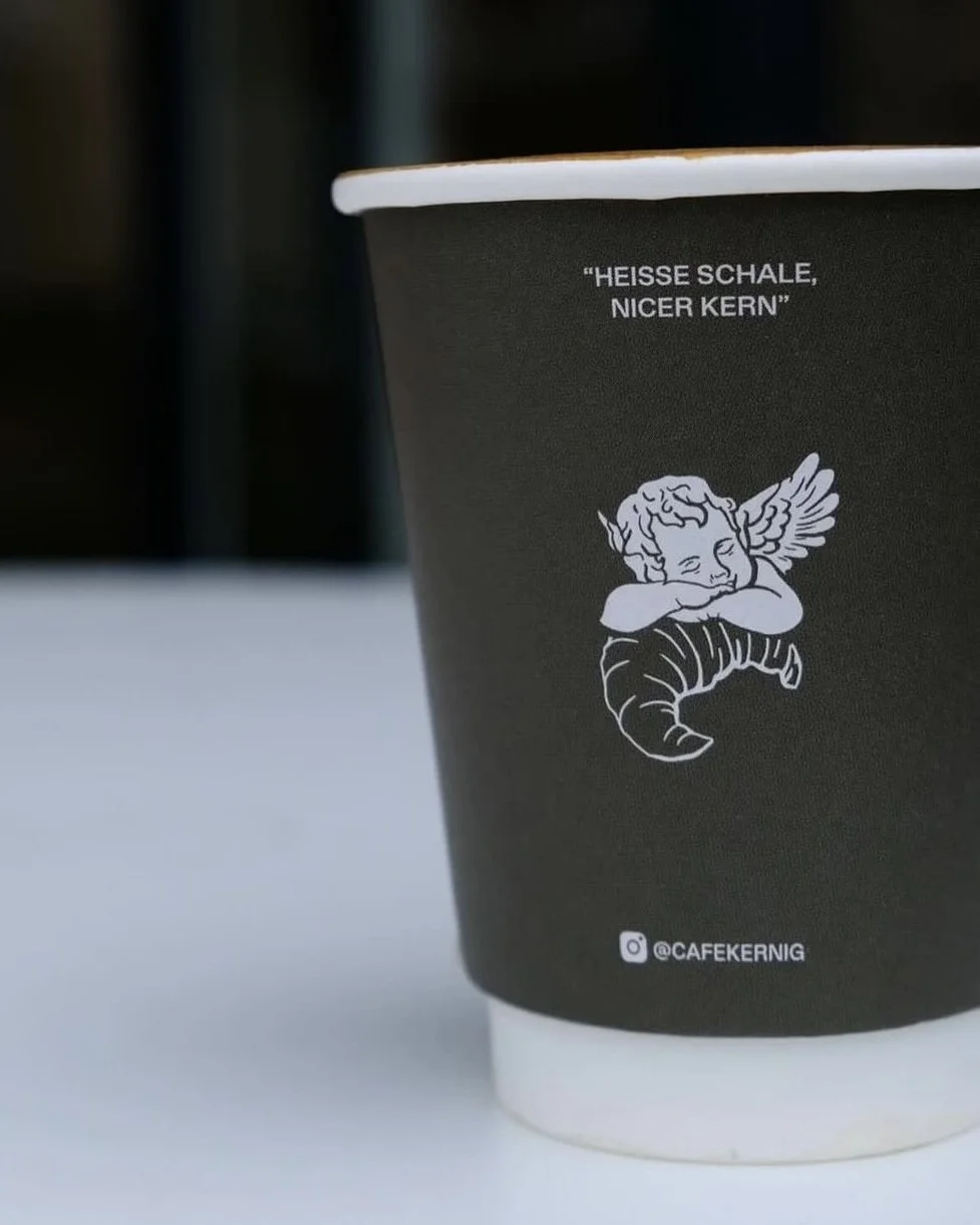 Schwarner Kaffeebecher mit Zeichnung eines schlafenden Engels und deutschem Text "Heisse Schale, Nicer Kern" sowie Instagram-Handle @CAFEKERNIG.