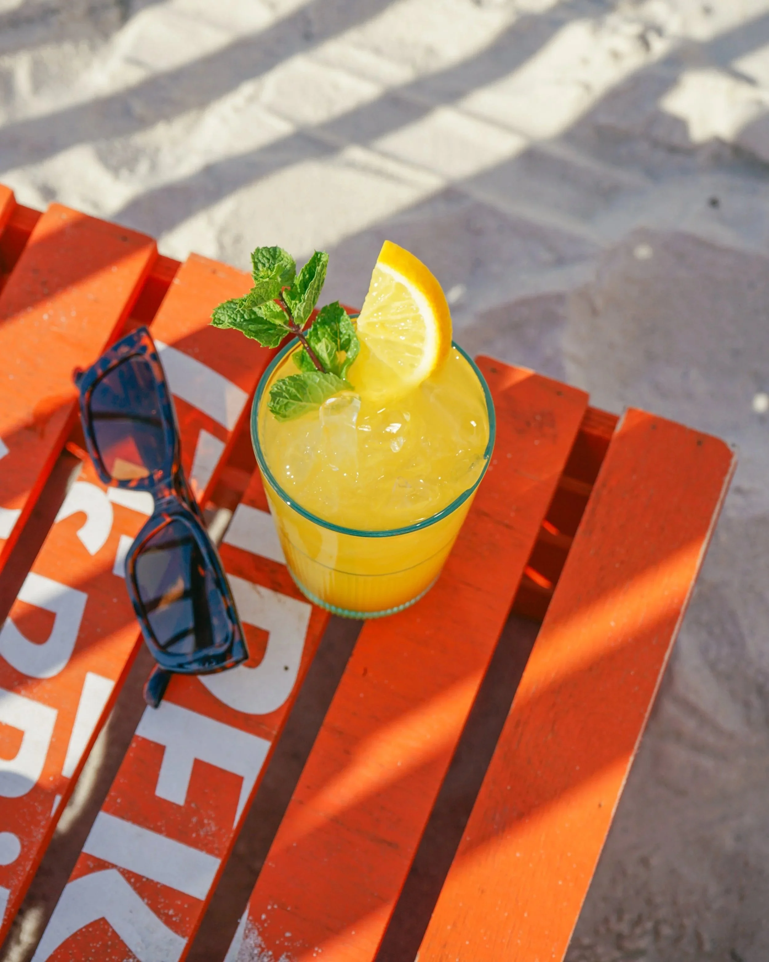 Ein Glas mit Zitronenlimonade, Zitronenscheibe und Minzblatt auf einem orangefarbenen Tisch am Strand.
