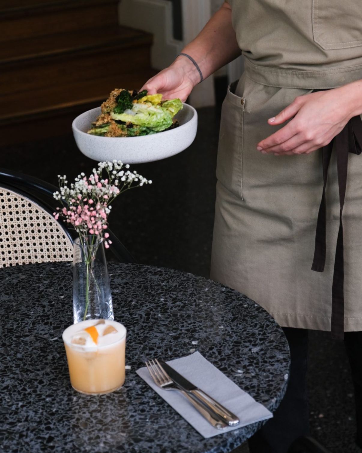 Ein Kellner hält einen Teller mit einem Salat. Im Vordergrund ist ein Tisch mit einer Vase mit rosa Blumen, einer Kerze, Besteck und einem Papierserviette.