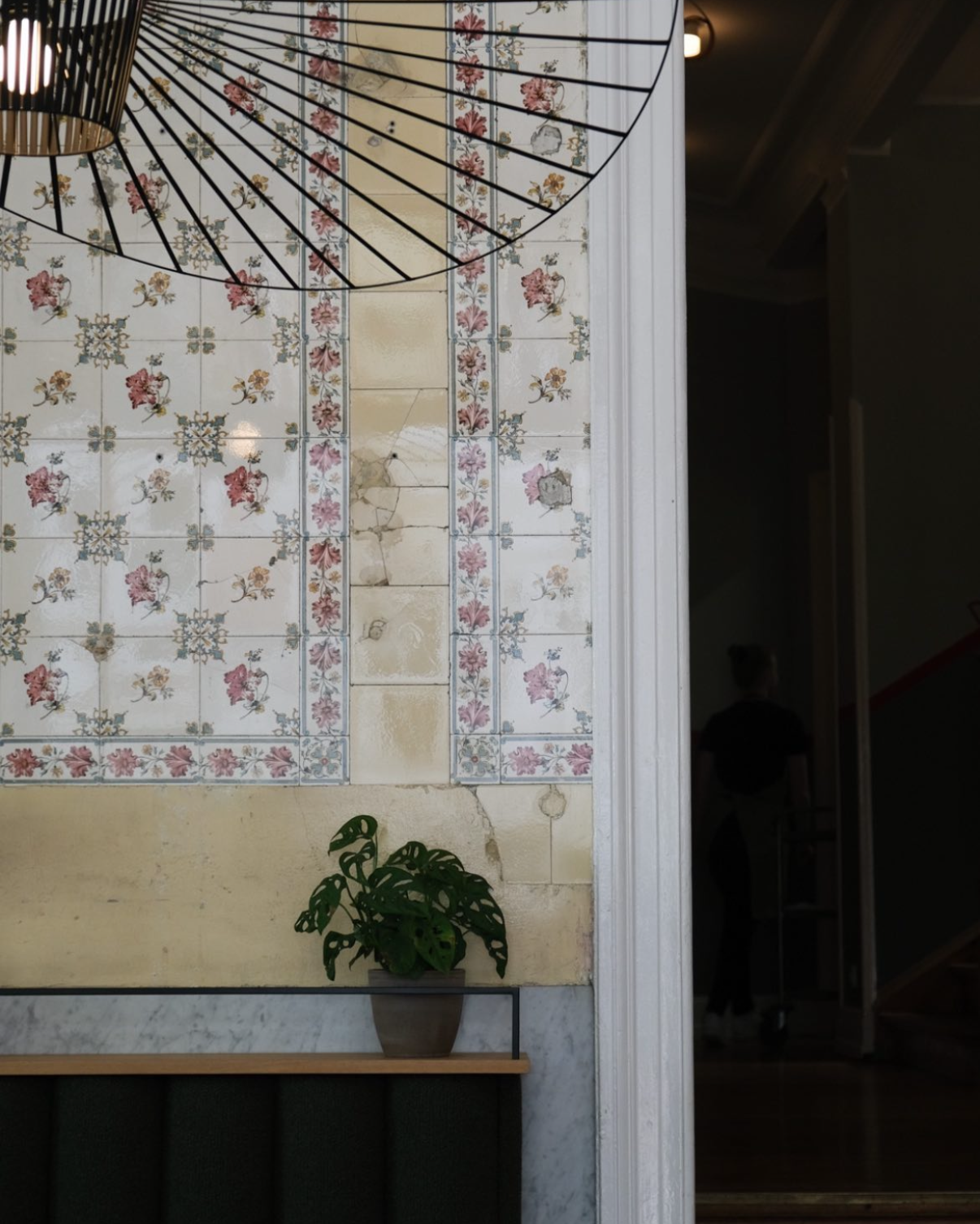 Blick auf eine Wand mit alten, abgetretenen Fliesen, darunter ein PflanzenTopf mit Grünpflanze auf einem Regal, rechts eine weiße Wandverkleidung.