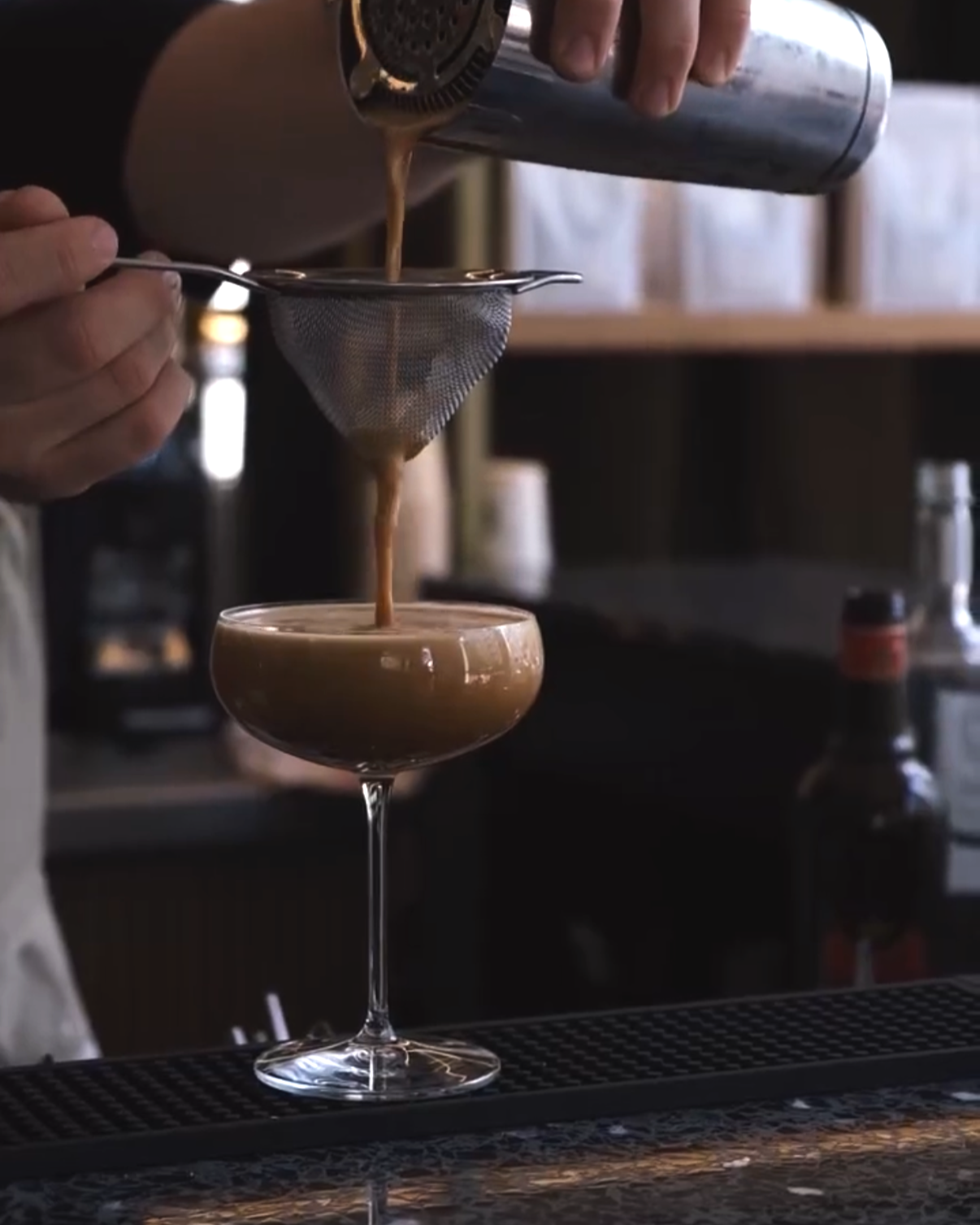 Jemand gießt Kaffee durch ein Sieb in ein Cocktailglas auf einem Bar-Tresen.