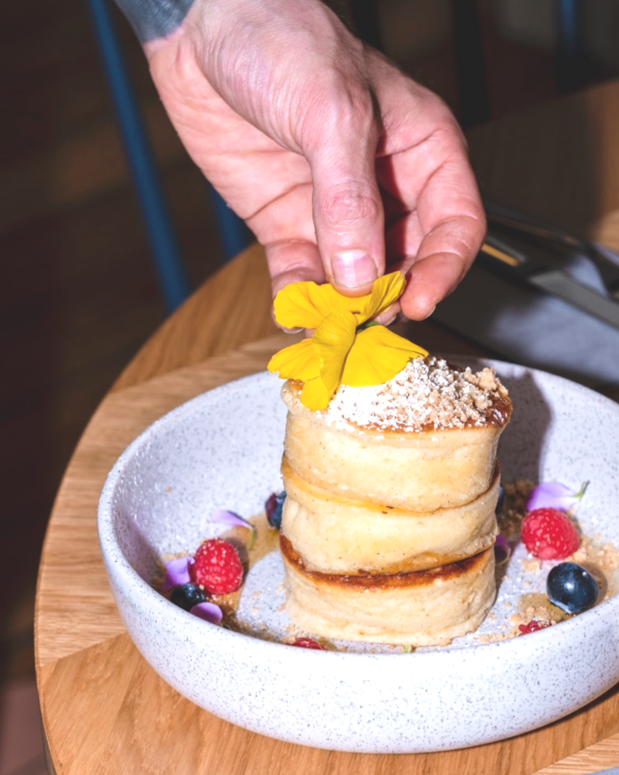 Hand, das ein Dessert mit Blumen und Beeren auf einem weißen Teller auf einem runden Holztisch arrangiert.