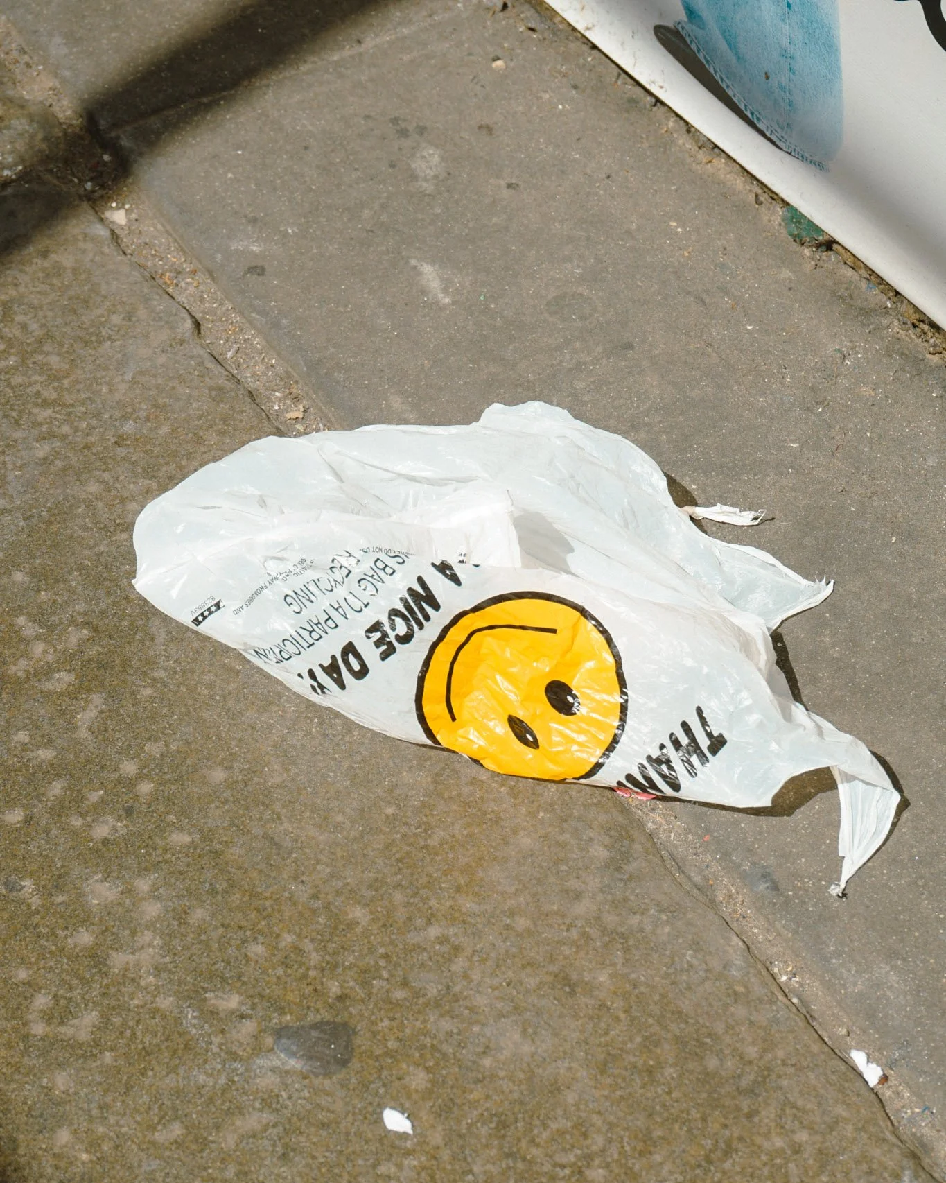 Eine weggeworfene Plastiktüte mit einem lachenden Smiley und Text darauf, liegt auf dem Bürgersteig neben einer Straßenkante.
