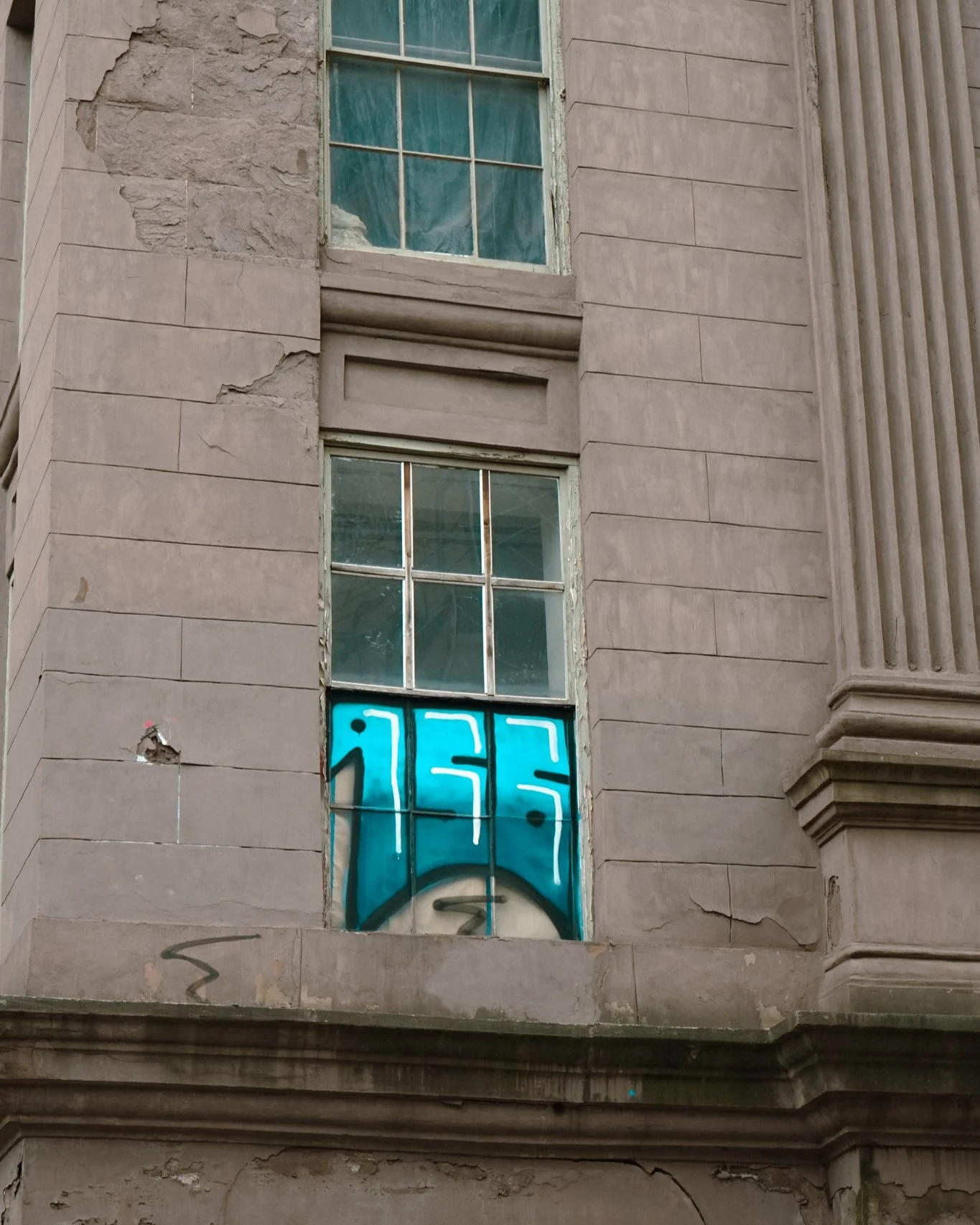 Ein altes, abgenutztes Gebäude mit kaputtem Mauerwerk und einer verfallenen Fensterfront. Im unteren Fenster ist Graffiti in Blau und Schwarz zu sehen.