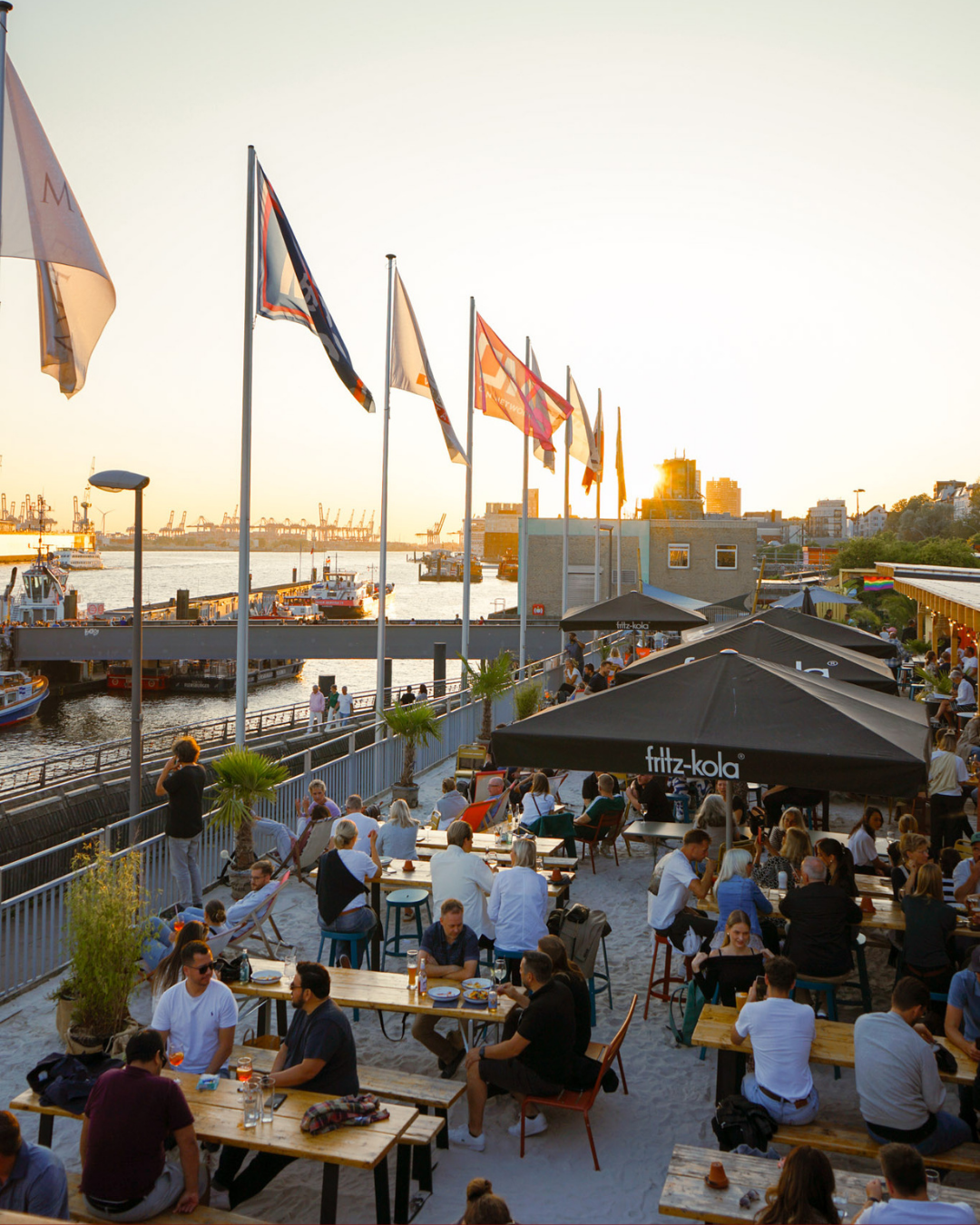 Menschen sitzen an Tischen in einem Outdoor-Restaurant am Wasser bei Sonnenuntergang, mit Flaggen auf hohen Masten und Booten im Hintergrund.