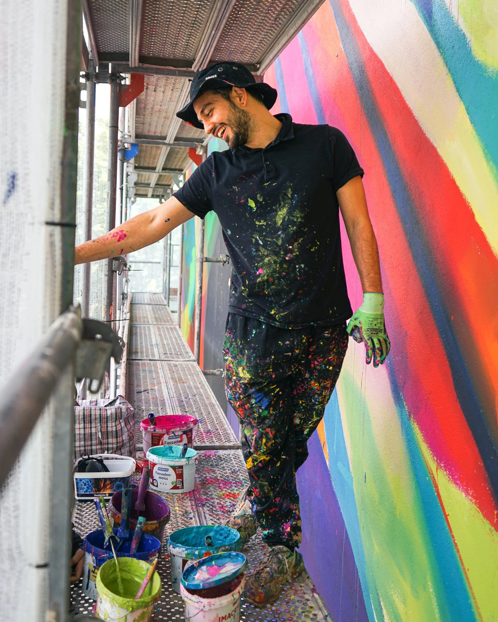 Ein junger Mann, gekleidet in eine schwarze Kleidung mit Farbspritzern, arbeitet an einer bunten Wandmalerei. Er trägt einen schwarzen Hut, hat einen Bart und lächelt glücklich, während er an einem Gerüst steht, um die Wand zu bemalen.