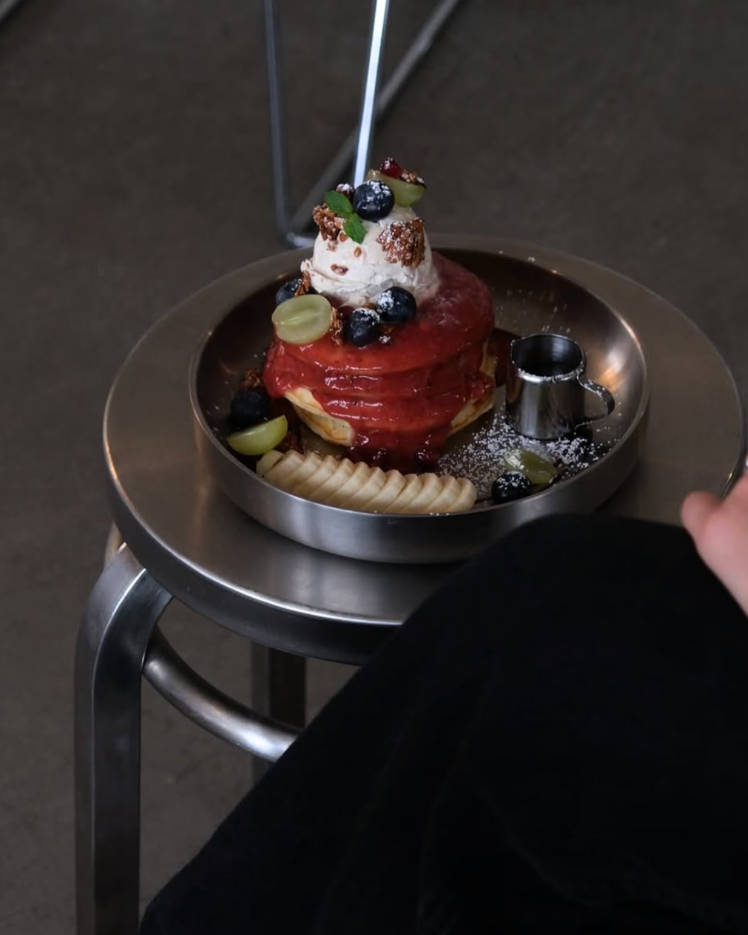 Ein Teller mit einem Pfannkuchen, bedeckt mit roter Fruchtsoße, verziert mit Heidelbeeren, grünen Trauben, einem Klecks Schlagsahne und Krokant. Ein kleines Messbecher-Behälter ist ebenfalls auf dem Teller. Der Teller liegt auf einem kleinen, runden,