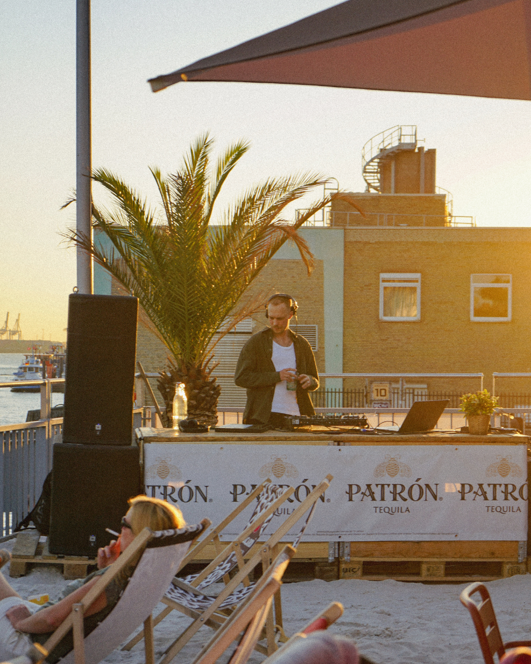 DJ spielt Musik an einem Strand bei Sonnenuntergang, während entspannte Menschen in Liegestühlen sitzen, im Hintergrund Gebäude und ein Palmbaum.