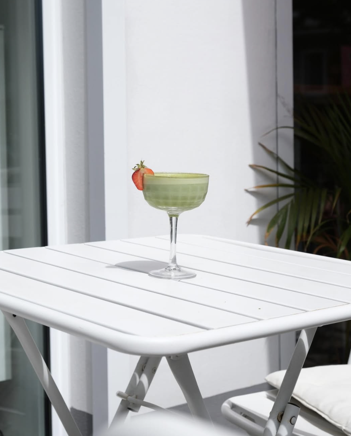 Ein grüner Drink in einem Cocktailglas mit einer Erdbeere als Garnierung auf einem weißen Balkon oder Terrasse.