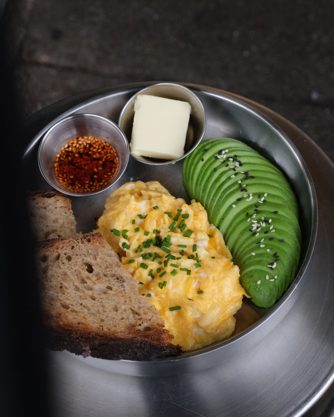 Frühstücksteller mit Avocado, Rührei, Vollkornbrot, Butter, und Sojasauce auf einem Metalltablett.