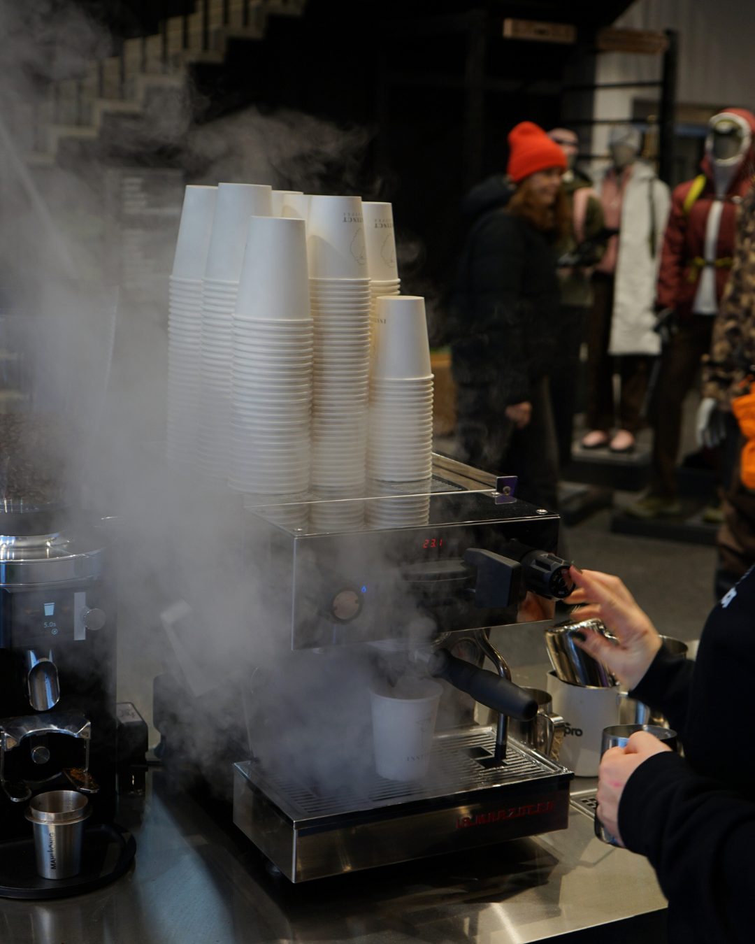 Menschen beim Kaffeebrühen mit Espressomaschine in einem Café, im Hintergrund wartende Gäste in Winterkleidung.