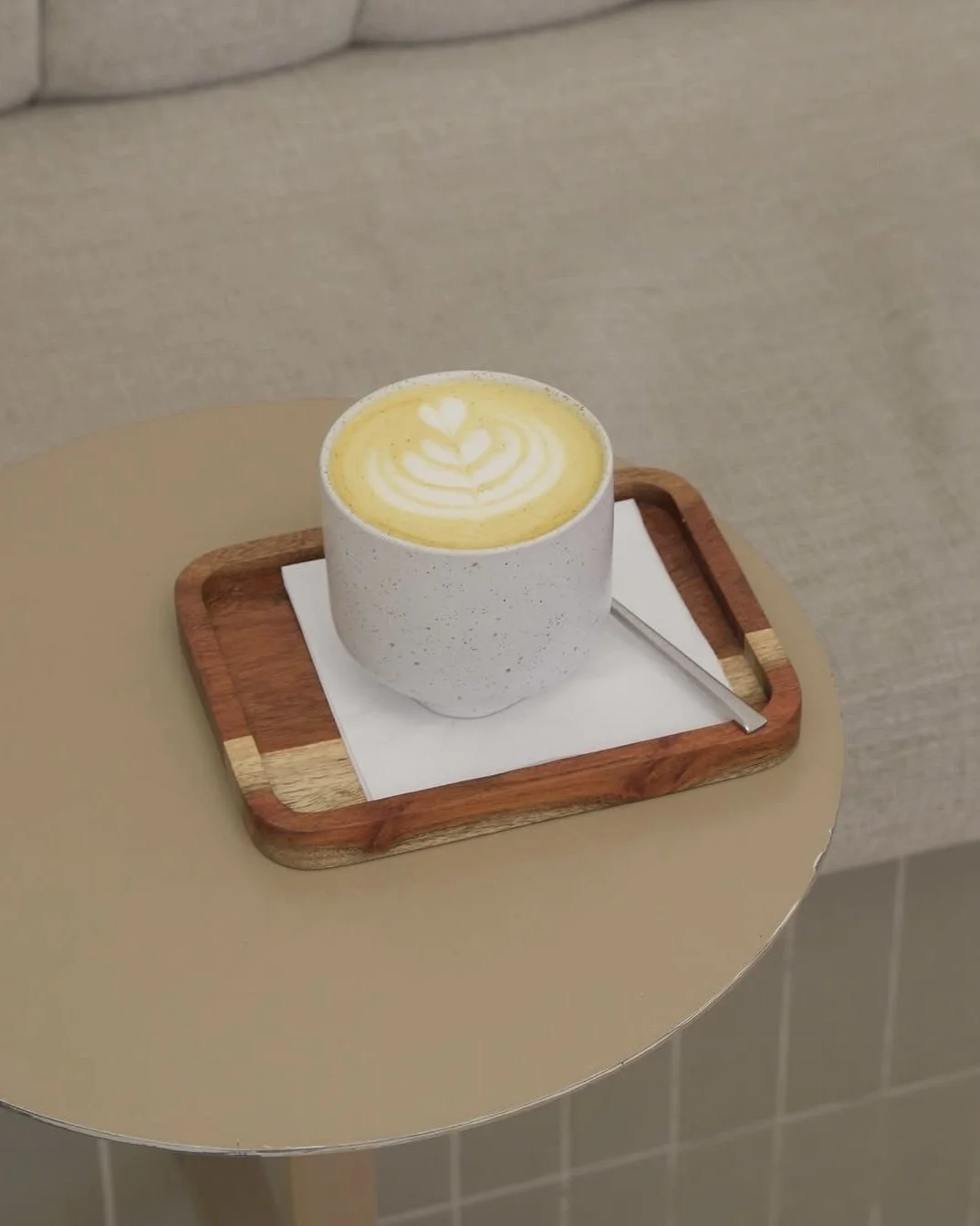 Eine weiße Tasse mit Kaffee oder Latte auf einem Holztablett, das auf einem runden beige Tisch steht.