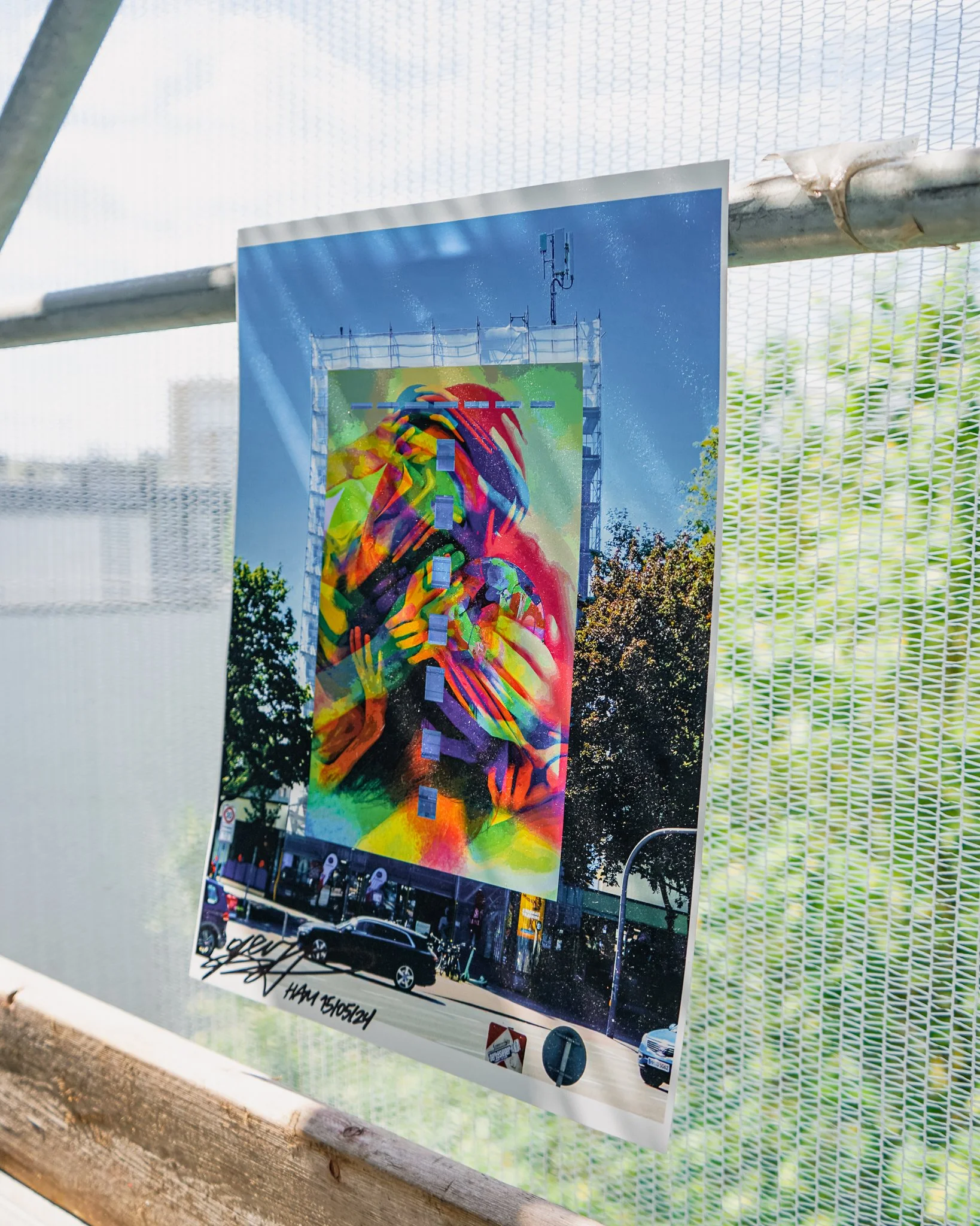 Ein farbenfrohes Wandbild eines stilisierten Menschen mit langen Haaren, der in die Hände seines Kopfes greift, ist an einem Baugerüst befestigt. Dahinter sind Bäume und eine Straße mit Autos sichtbar.
