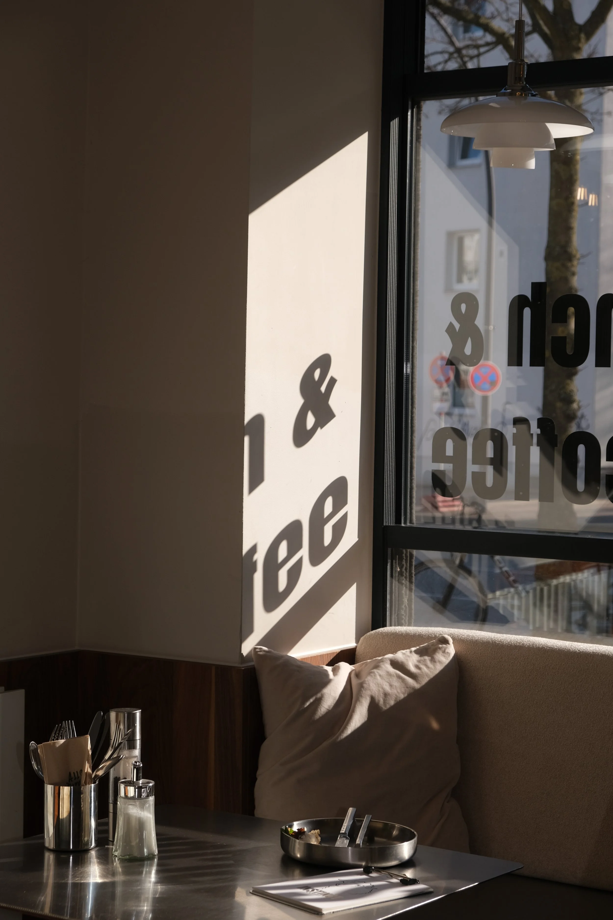 Ein sonnendurchflortes Café mit einem großen Fenster, das Schatten von Zeitschriften oder Werbung auf die Wand wirft. Im Vordergrund eine Tafel mit Besteck, Salz und Pfeffer, eine Schachtel mit Zigarette und eine Zeitung, eine Couch mit Kissen.