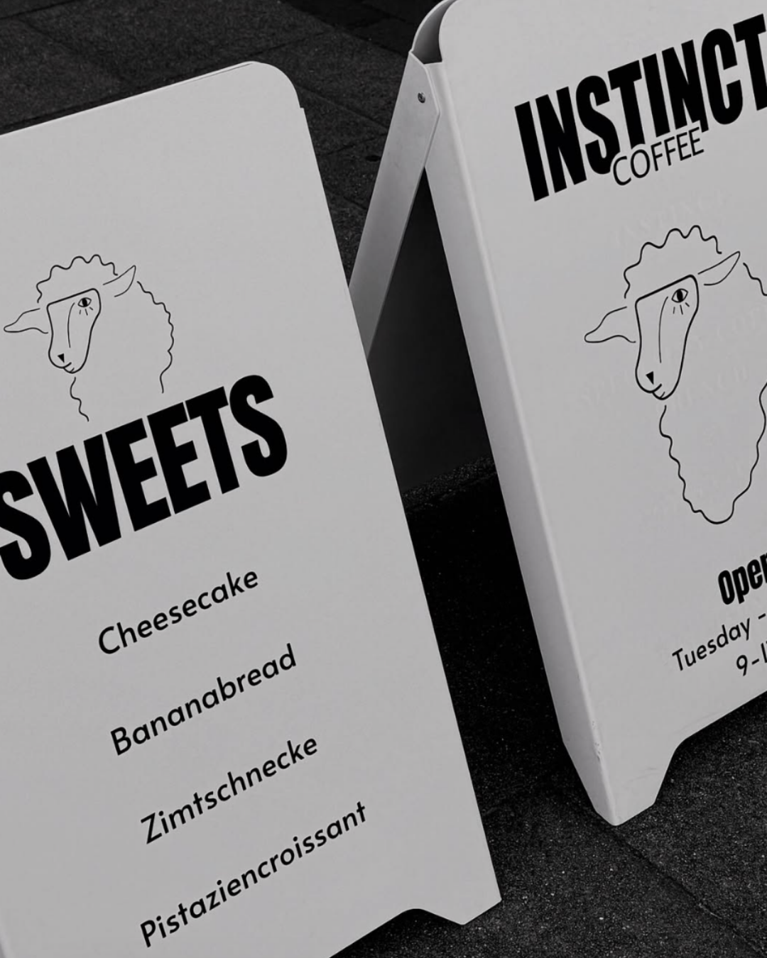 Zwei weiße Werbetafeln für ein Cafe namens 'Instance Coffee', mit einem einfachen schwarzen Lamm-Zeichnung und Menüangeboten wie Cheesecake, Bananabread, Zimtschnecke, Pistaziencroissant.