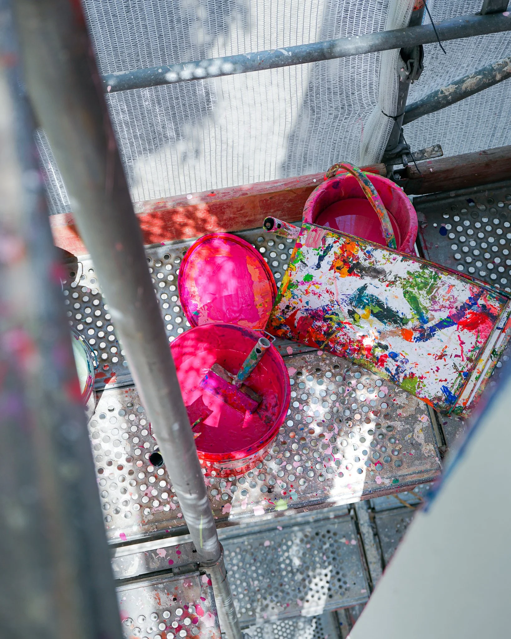 Bunte Eimer und eine Malpalette auf einer hölzernen und metallenen Plattform, die mit Farbspritzern bedeckt sind.