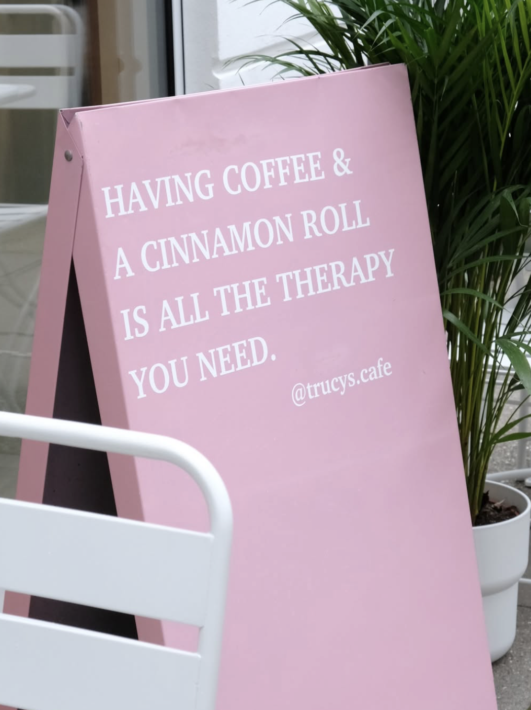 Ein pinkfarbiger Sandwich-Board-Schilder mit dem Text: 'HAVING COFFEE & A CINNAMON ROLL IS ALL THE THERAPY YOU NEED. @trucys.cafe', steht vor einem grünen Pflanzen.