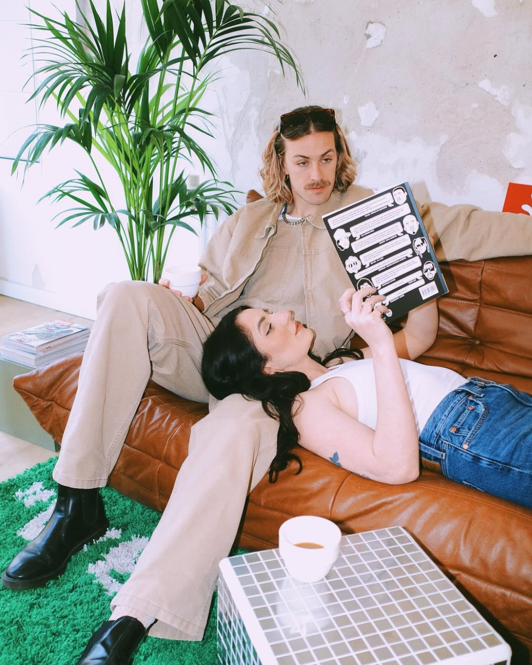 Ein Mann und eine Frau entspannen sich auf einem braunen Ledersofa. Der Mann sitzt mit einem Buch, die Frau liegt mit dem Kopf auf seinem Schoß. Es gibt eine grüne Pflanze im Hintergrund und eine Tasse Kaffee auf einem Tisch vor ihnen.