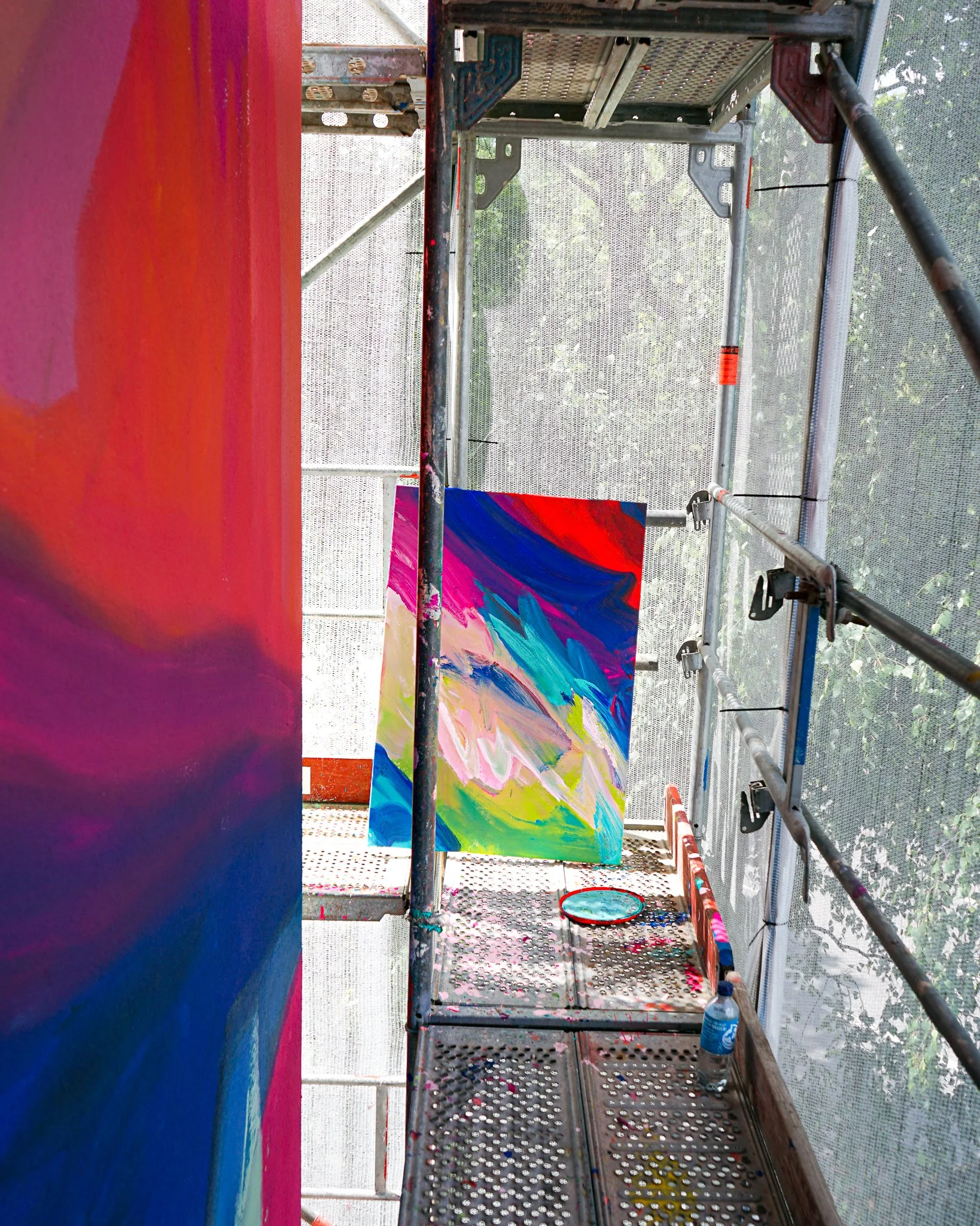 Ein farbenfrohes abstraktes Gemälde in einem Kunststudio, das auf einer Mauer angebracht ist, mit einem Malerei-Setup auf einer Metallplattform, Pinsel und Farbspachtel auf dem Boden und eine Wasserflasche.