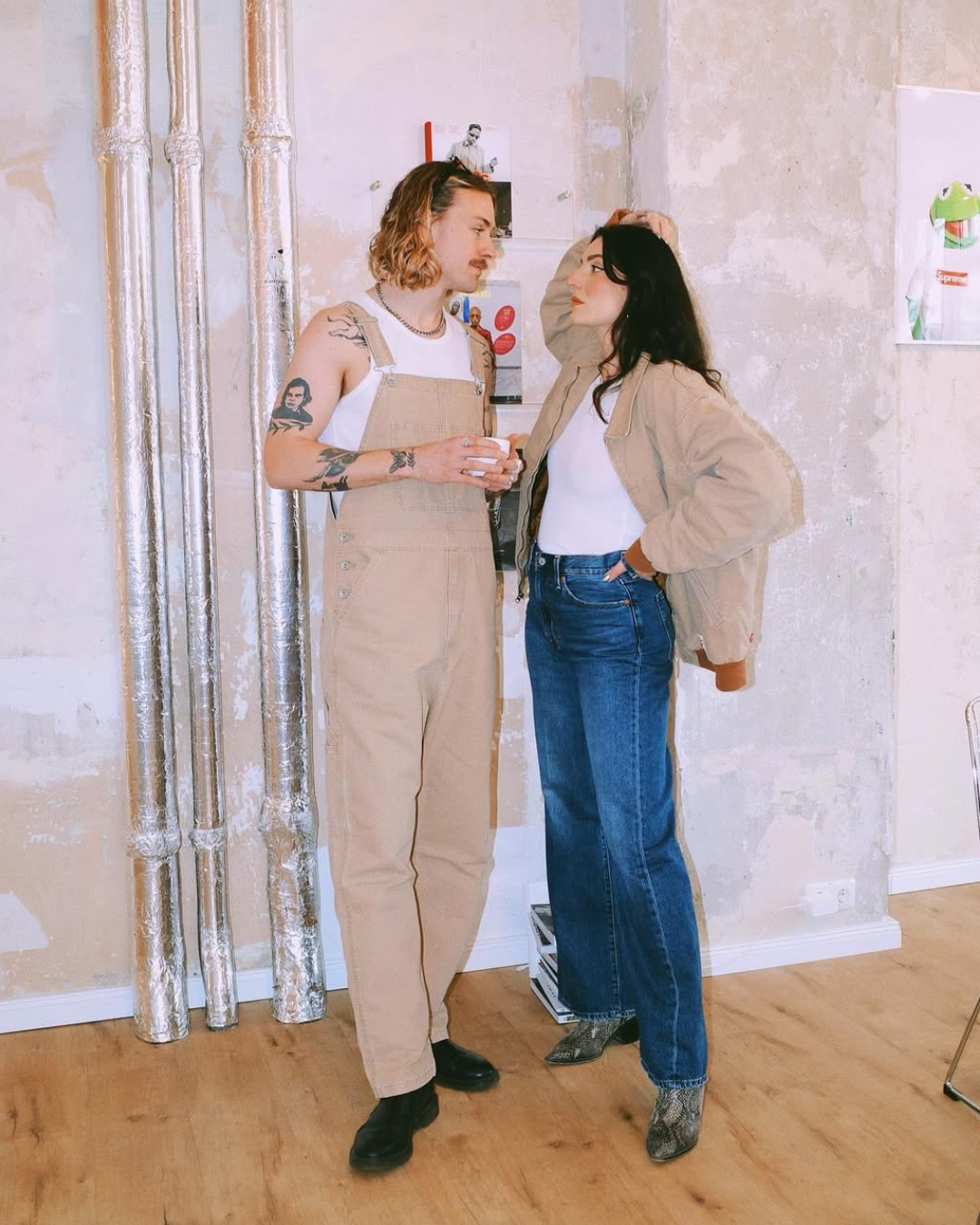 Zwei Menschen stehen sich gegenüber in einem Raum mit unvollendeter Wand. Der Mann trägt beige Latzhosen und hat Tattoos, die Frau trägt blue Jeans und eine beige Jacke. Sie schauen sich in die Augen, die Frau hat die Hand im Haar.