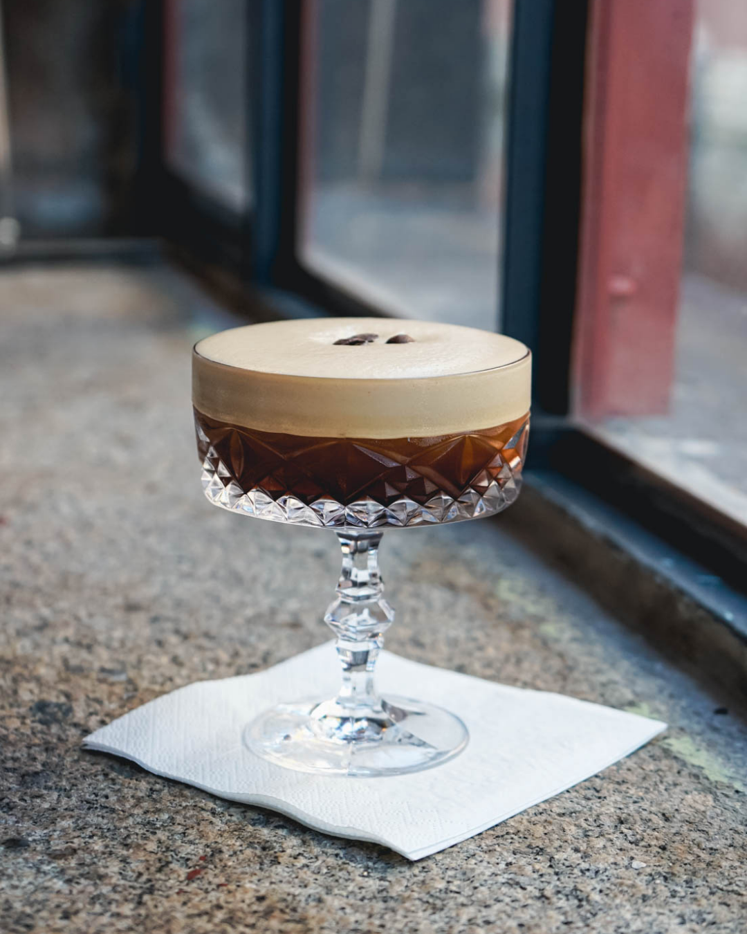 Ein koffeinhaltiges Getränk, wahrscheinlich Kaffee mit schaumiger Oberfläche, in einem dekorativen Kristallglas auf einer weißen Serviette am Fenster.