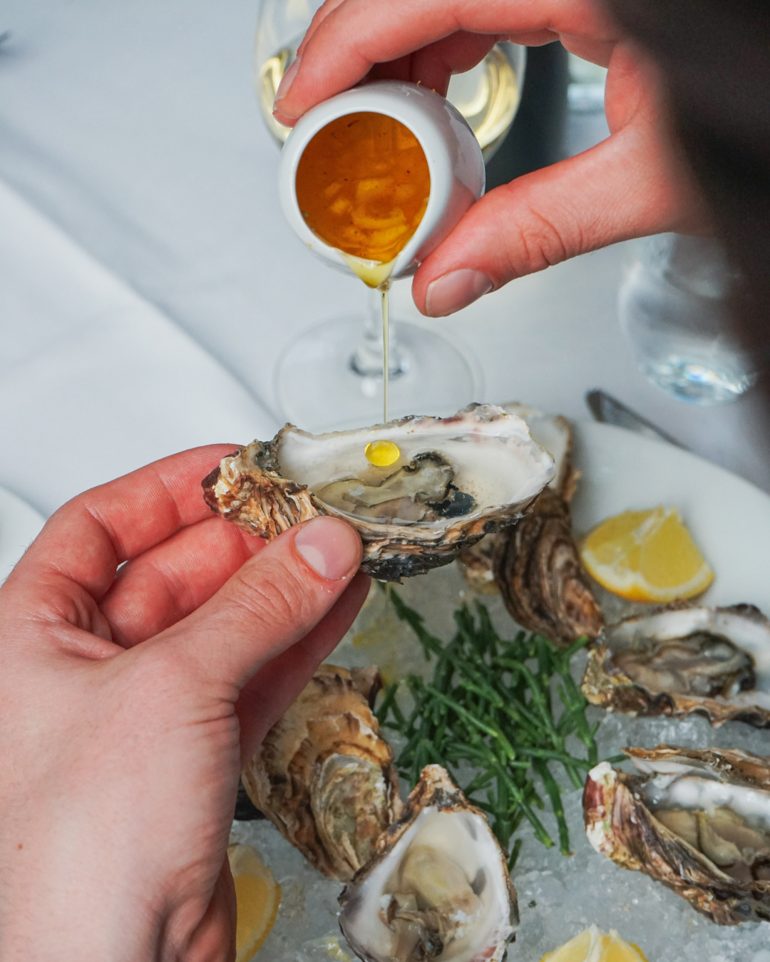 Person gießt Öl auf eine frische Auster, mit Meeresfrüchten, Zitronenscheiben und Kräutern auf dem Tisch.