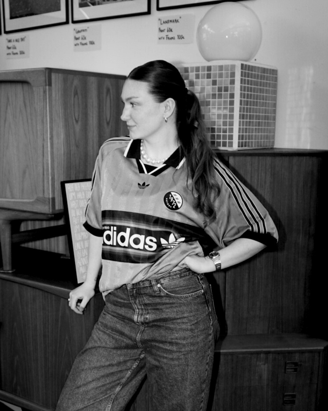 Junge Frau im Vintage-Stil in Sportbekleidung mit Adidas-Logo, schwarze Jeans, lässig lehnend in einem Raum mit Holzregalen und Wandbildern, schwarz-weiß Foto.