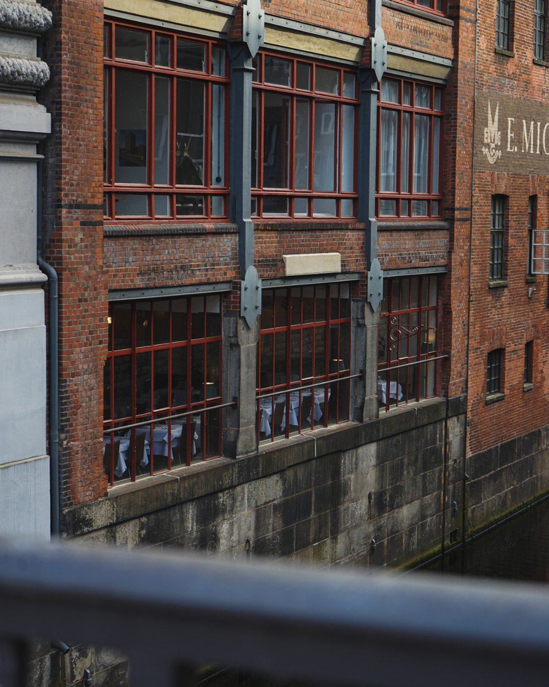 Ein altes Backsteingebäude mit großen, roten Fensterrahmen und Tischen mit weißen Tischtüchern im Erdgeschoss, das an einem Wasserweg liegt.