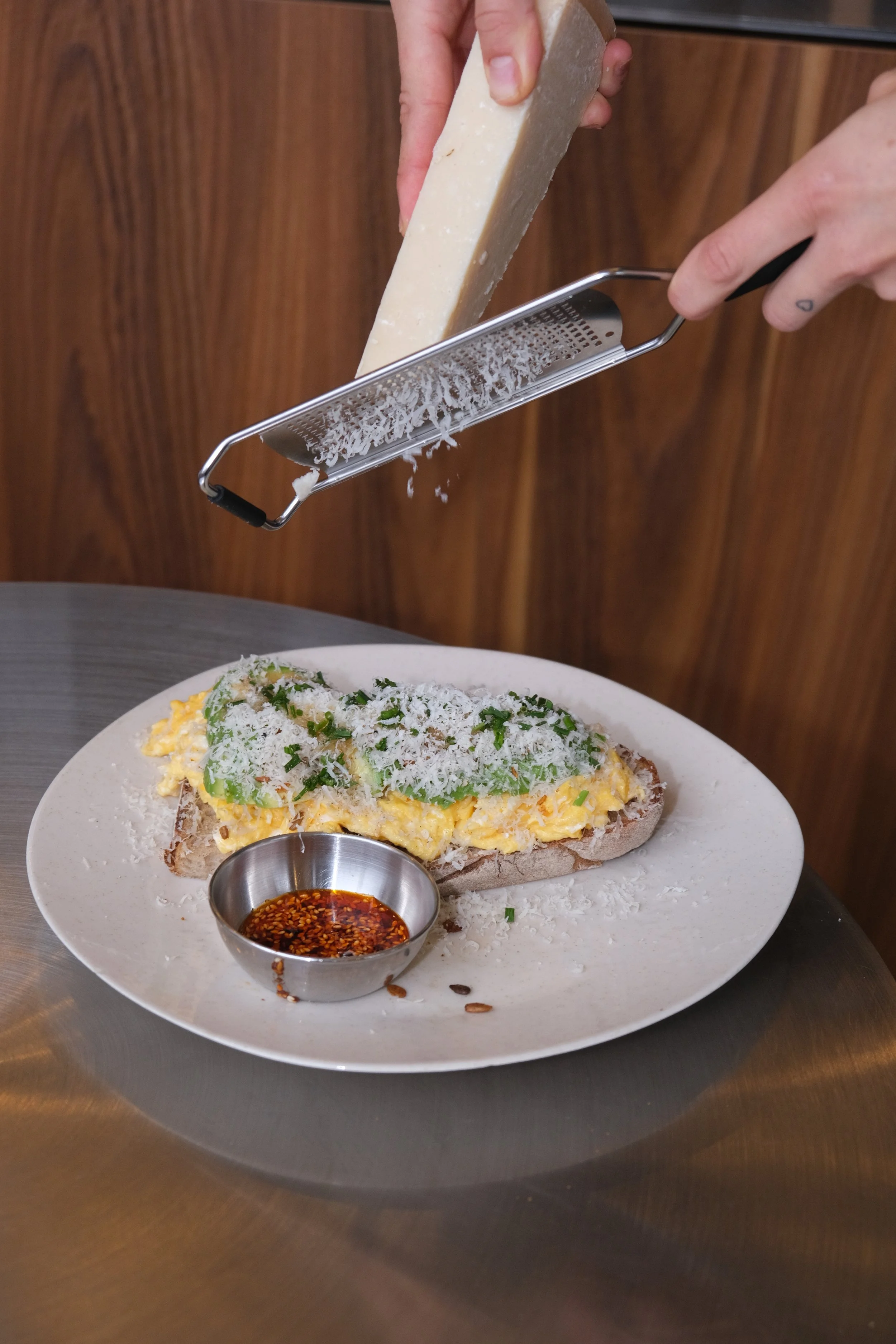 Hand reibt Käse über ein belegtes Brot mit Ei, Avocado und Kräutern, dazu eine kleine Schüssel mit Sojasauce oder Chiliöl.