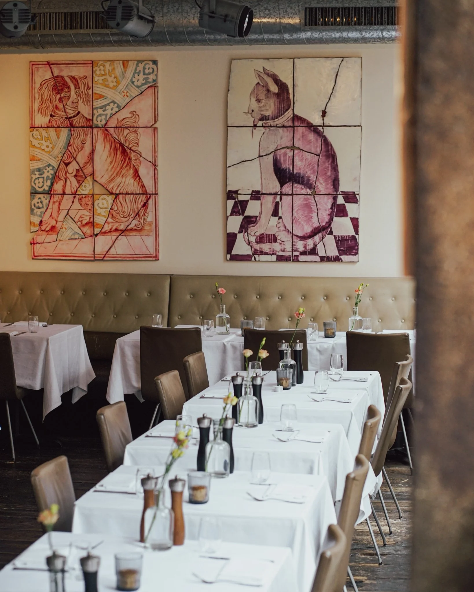 Ein schön gedeckter Restaurant-Tisch mit weißen Tischtüchern, Gläsern, Besteck und Vasen mit kleinen Blumen, im Hintergrund Wanddekoration mit zwei großformatigen, zerbrochenen Gemälden von Tieren.