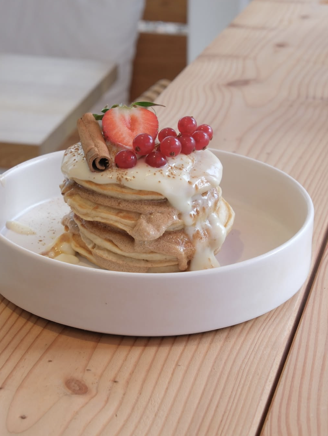 Pfannkuchen gestapelt mit Vanillesoße, garniert mit Erdbeere, roten Johannisbeeren und Zimtstange in einer weißen Schüssel auf Holzoberfläche.