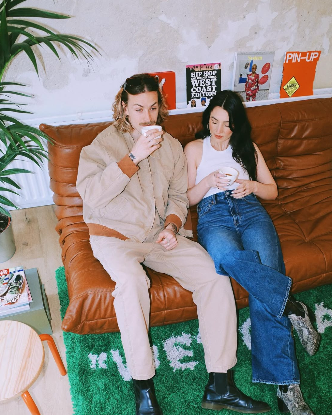 Zwei Menschen sitzen auf einem braunen Ledersofa, trinken Kaffee, in einem Raum mit Wanddeko und Büchern im Hintergrund.