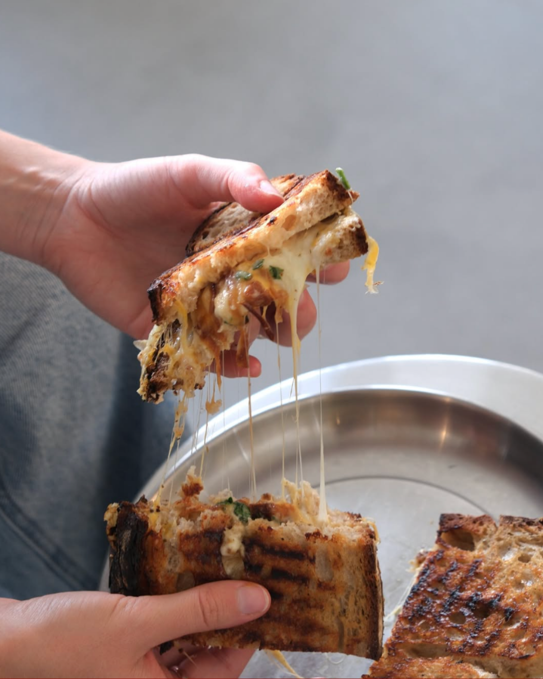 Nahaufnahme von zwei Händen, die eine knusprige Pizza oder belegtes Fladenbrot mit geschmolzenem Käse und Käsefäden hochheben, im Hintergrund ein runder Tablett.