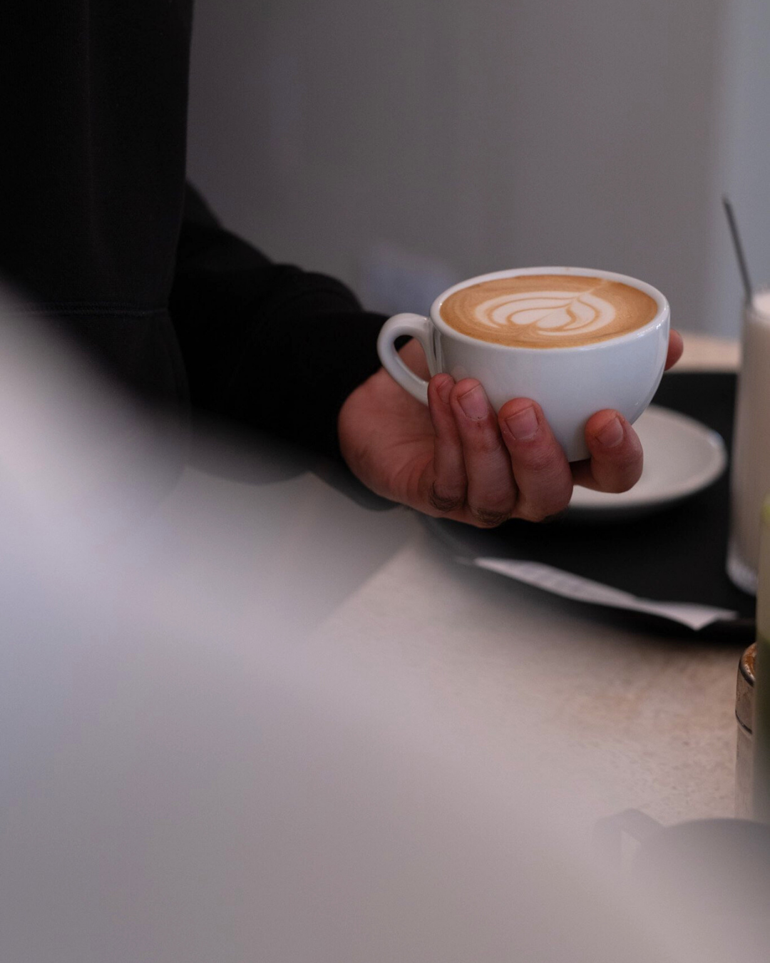 Eine Hand hält eine weiße Kaffeetasse mit einem Latte, das kunstvoll mit Milchschaum verziert ist.